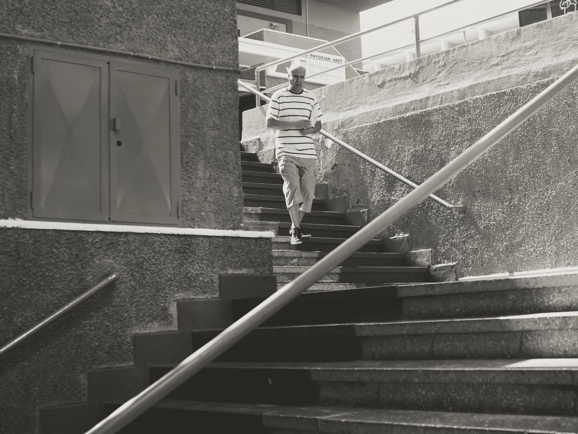 A man in a striped shirt and light-colored pants descending outdoor stairs with a metal handrail, with a rough-textured wall and building structure nearby.