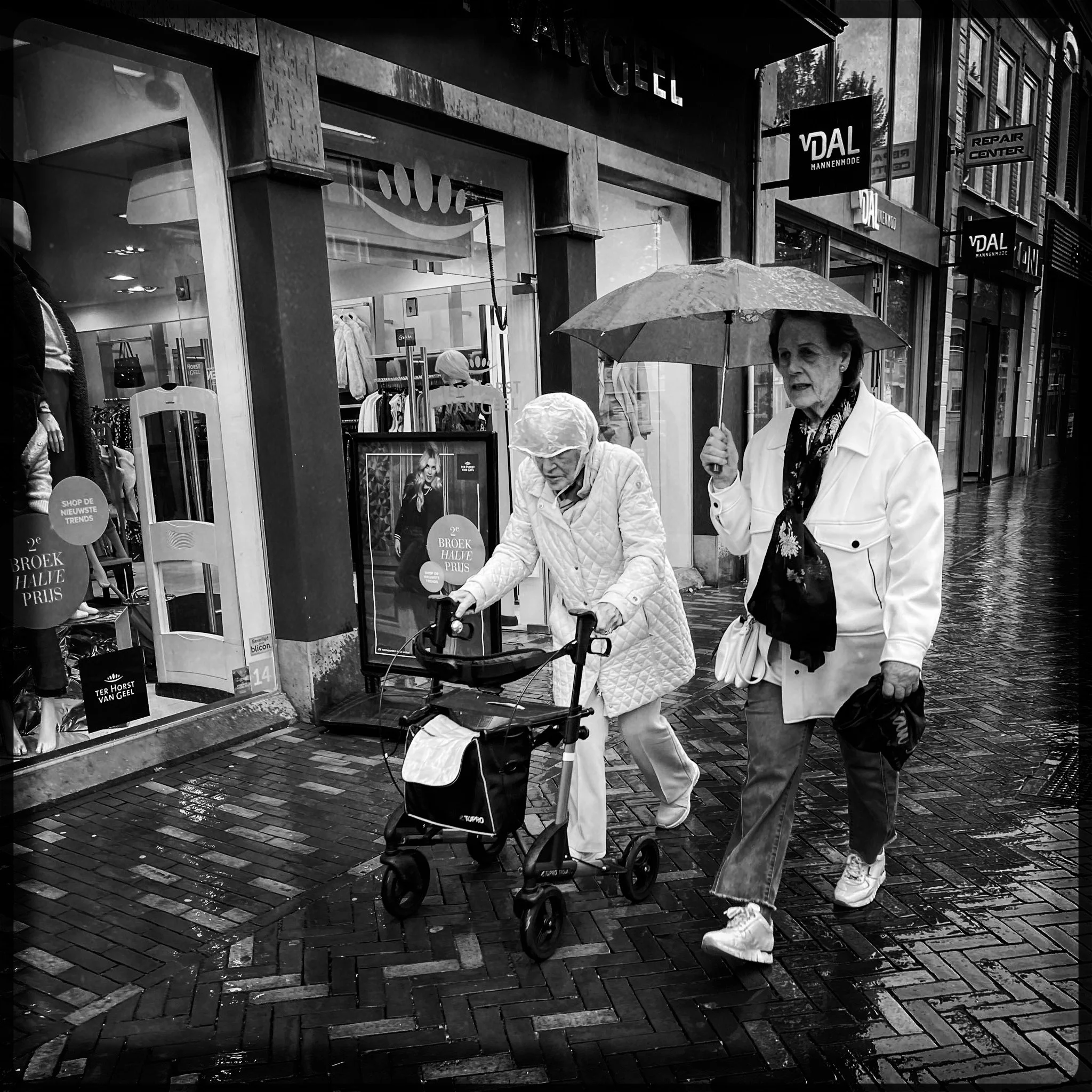 Two elderly women walking on a wet city street in the rain, one is using a walker and the other is holding an umbrella, both dressed in light-colored coats.