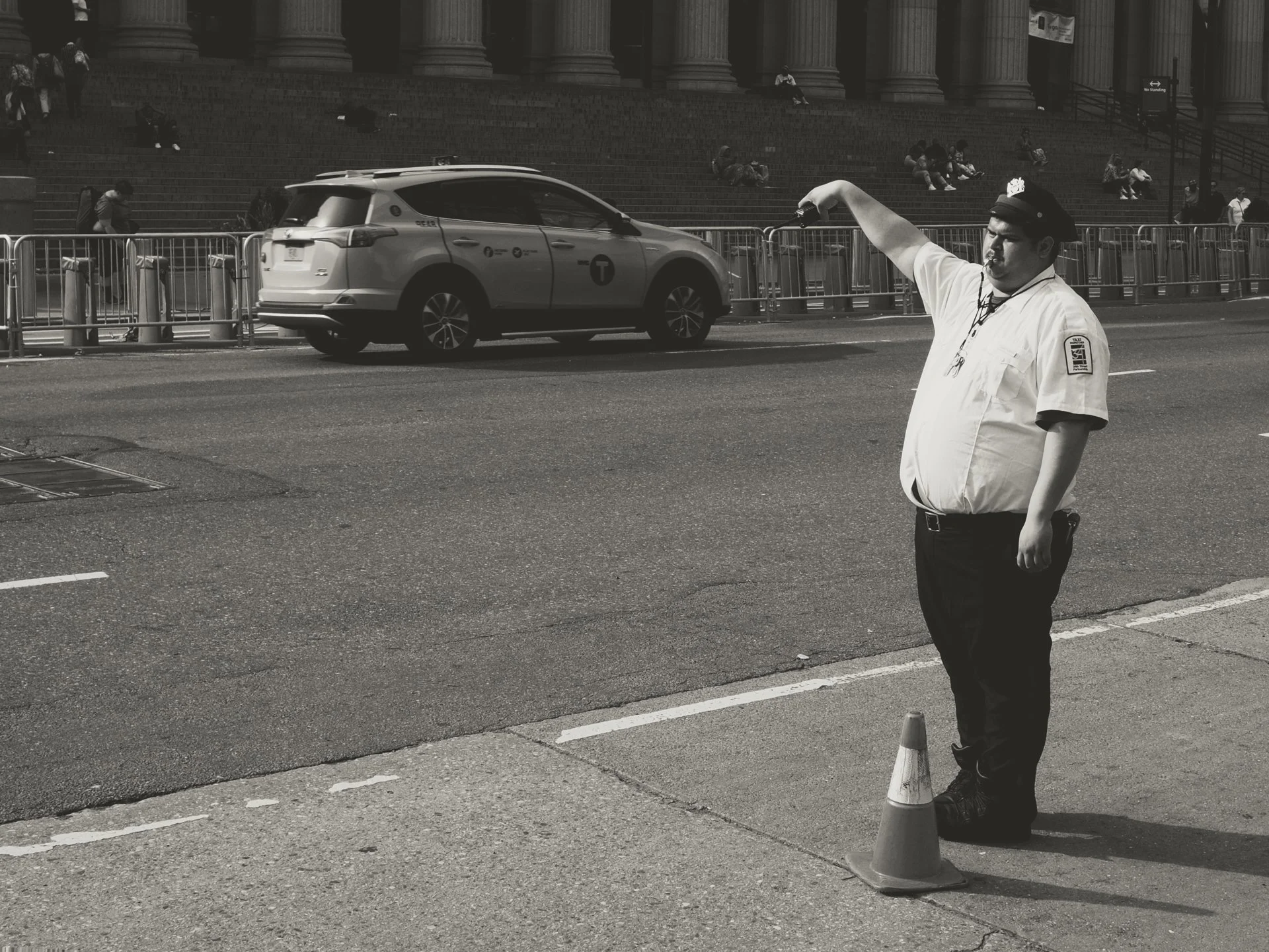 A traffic officer in uniform is directing traffic in front of a building with large pillars. The officer is standing next to traffic cones and appearing to give signals or instructions. Several people are sitting and standing on the steps of the buil