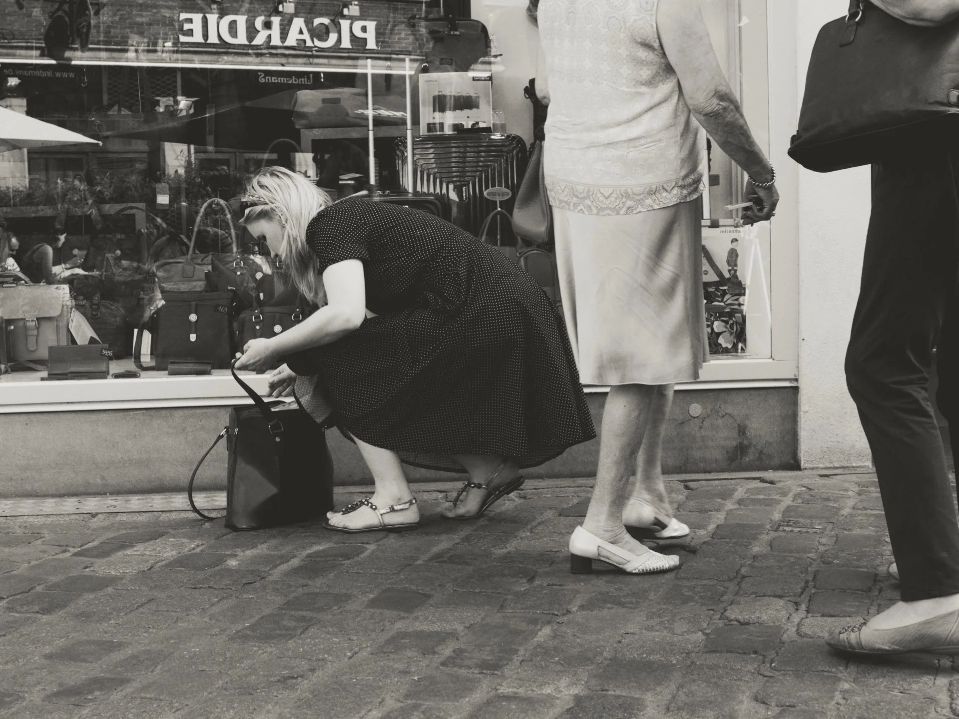 A woman wearing a polka dot dress crouches to look at items on display in a store window, with her purse on the ground beside her. Two other women are standing nearby, one with a handbag and the other holding a cigarette, all seen from the waist down