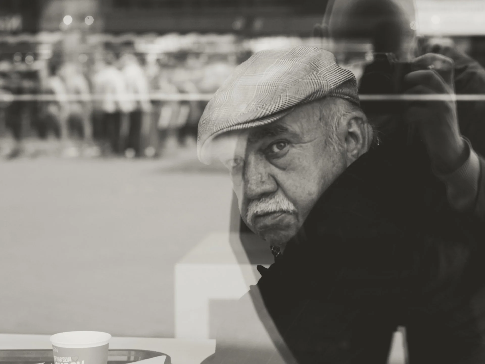 Older man with glasses and a checkered hat looking through a glass window at a McDonald's paper cup on a table.