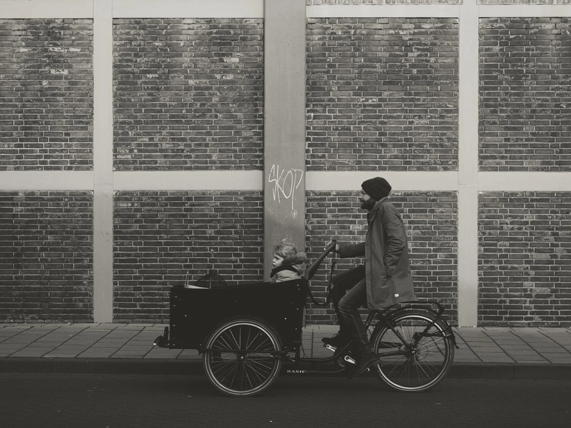 A man in a coat and beanie riding a bicycle with a child in a cargo bike on a city street. The background features a brick wall with repetitive patterns and a graffiti signature on a vertical concrete pillar.