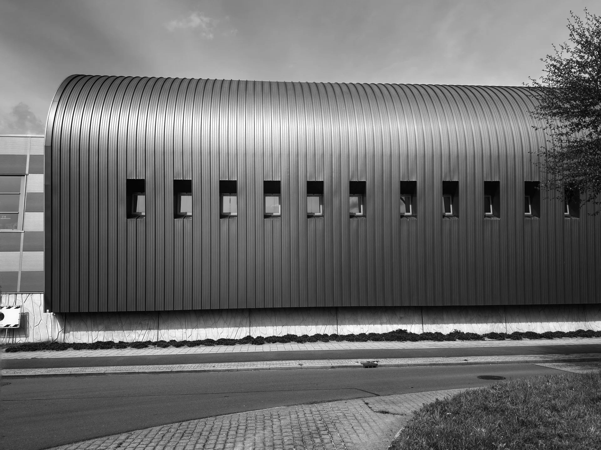 A modern building with a curved, metallic exterior and small rectangular windows, situated along a street with a sidewalk and grass.
