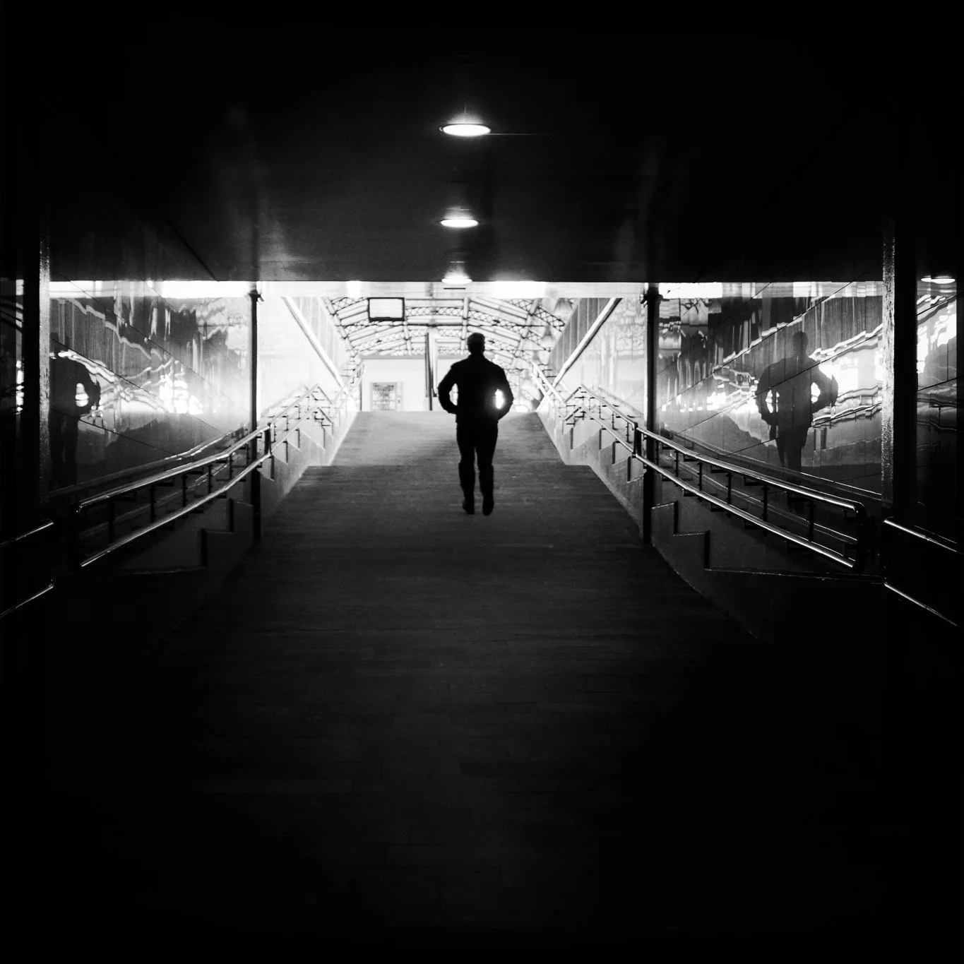 Silhouette of a man walking up a covered outdoor staircase with reflective walls on either side, leading to a bright exit.