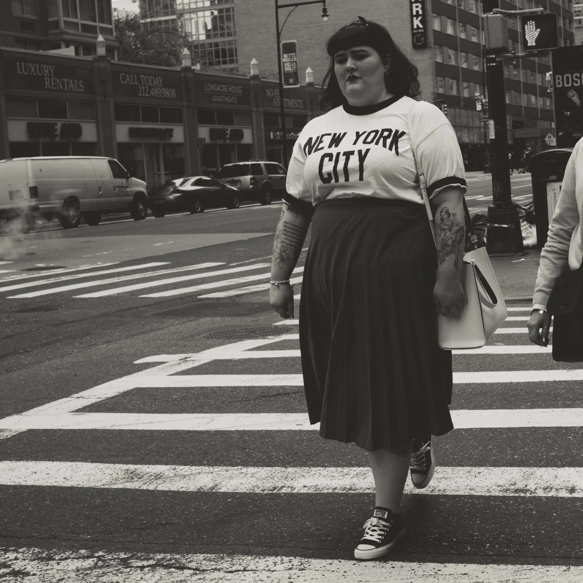 A person with dark hair and tattoos on their arm, wearing a white t-shirt with 'New York City' written on it, walking across a city street with cars and tall buildings in the background. The person is wearing sneakers and carrying a bag.
