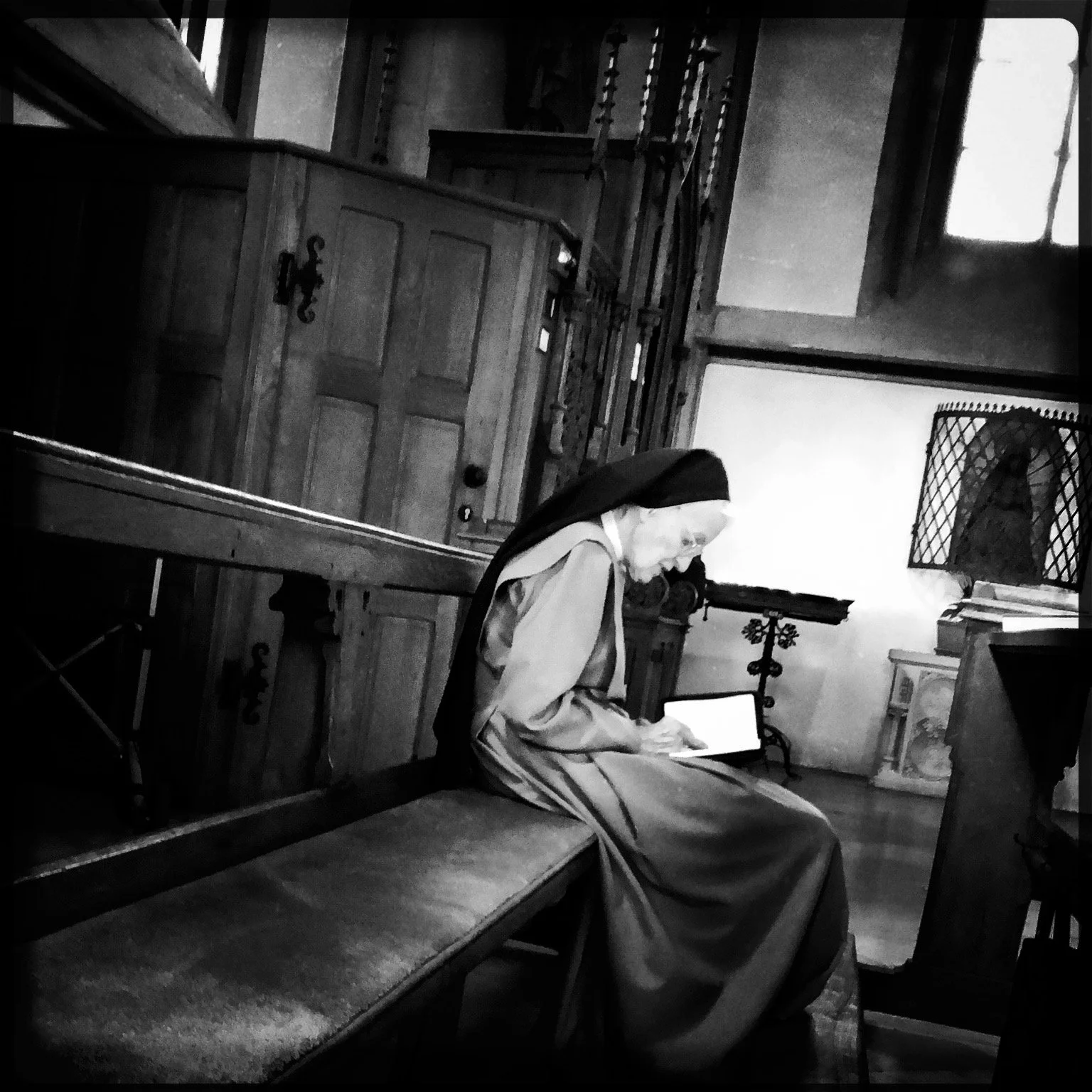 A nun in religious attire sitting on a bench reading a tablet inside a church or chapel.