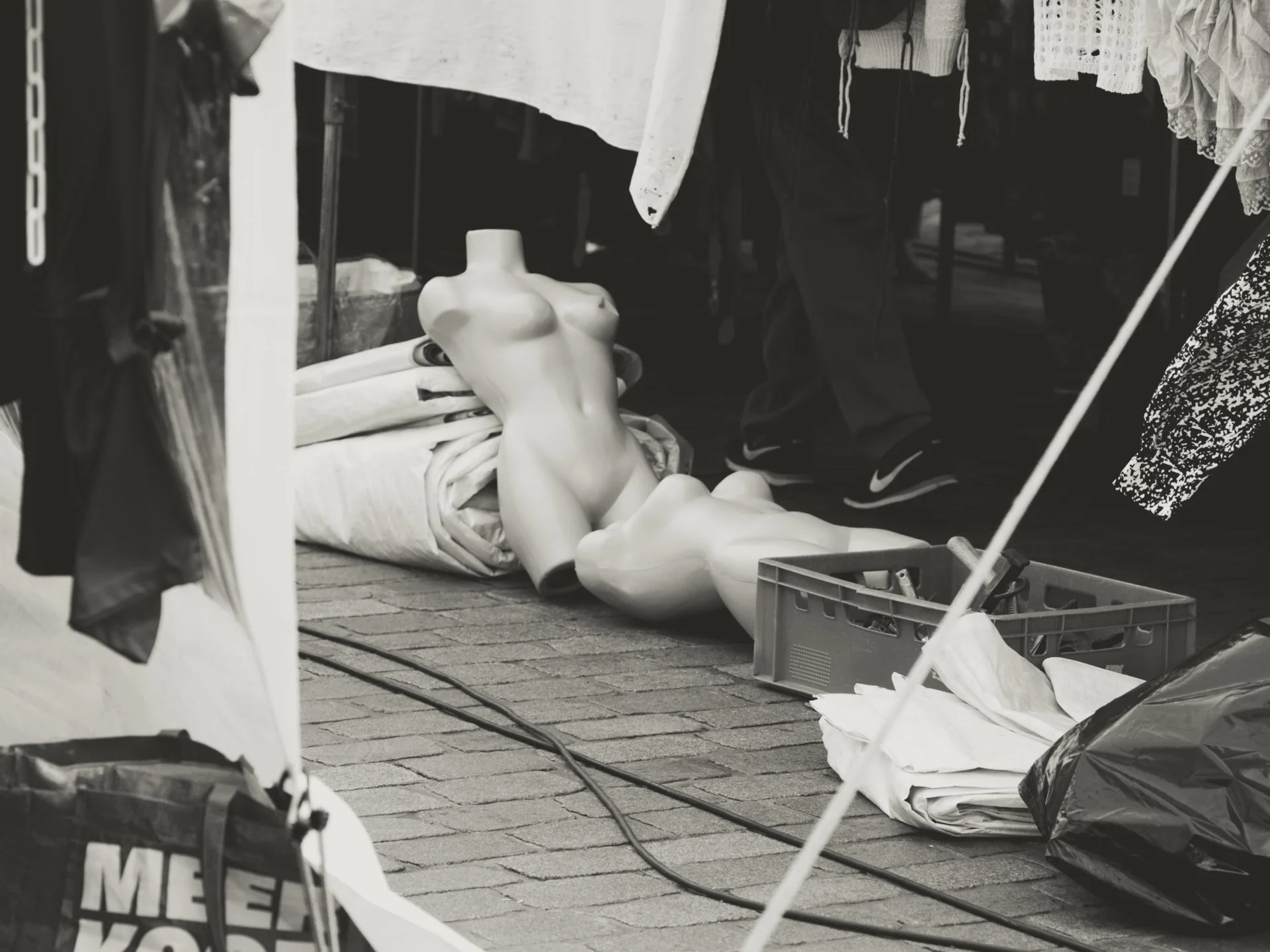 Street sale display with a female torso mannequin lying on the ground, surrounded by clothes, umbrellas, and shopping bags.