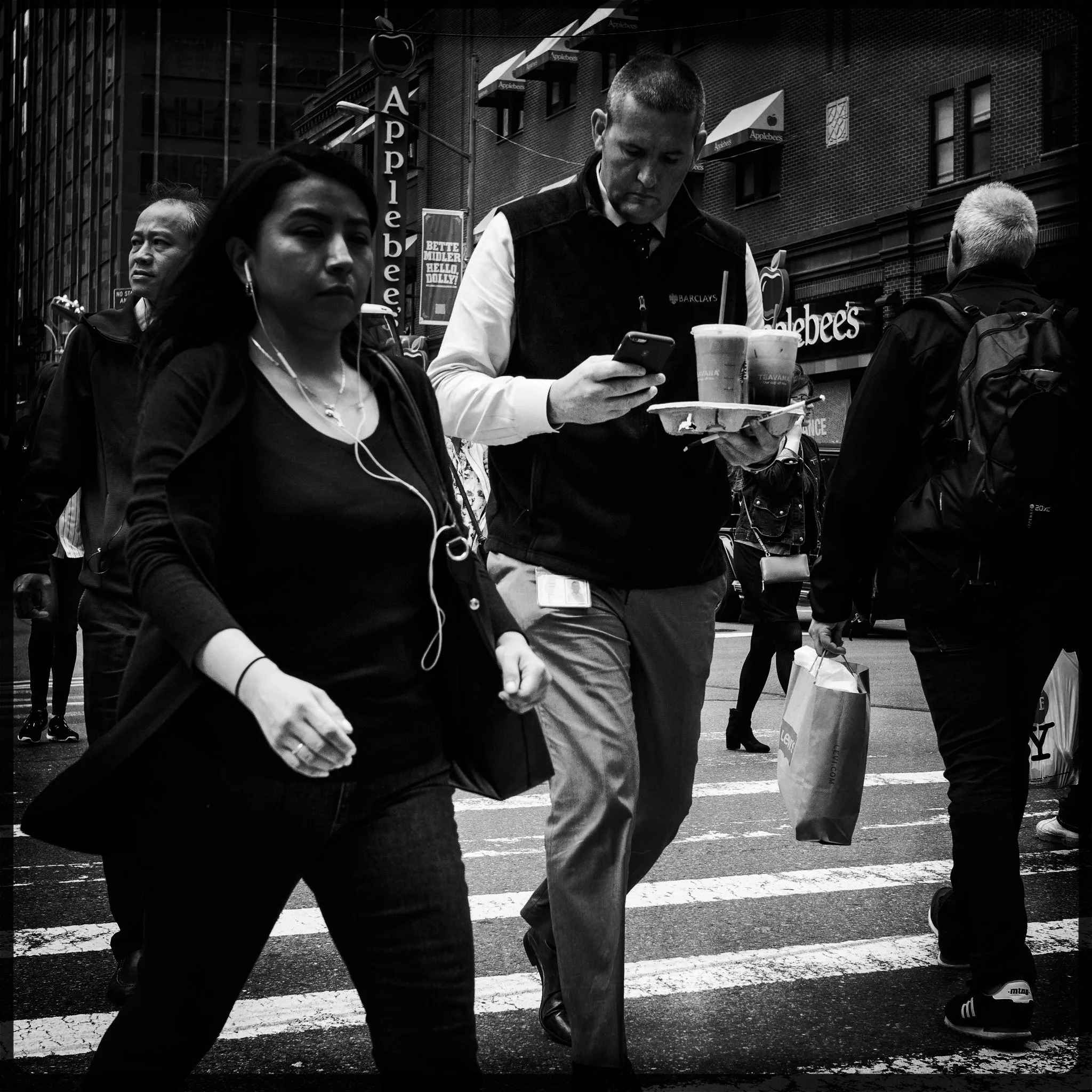 Black and white photo of pedestrians crossing a busy city street, with storefronts and signs in the background.