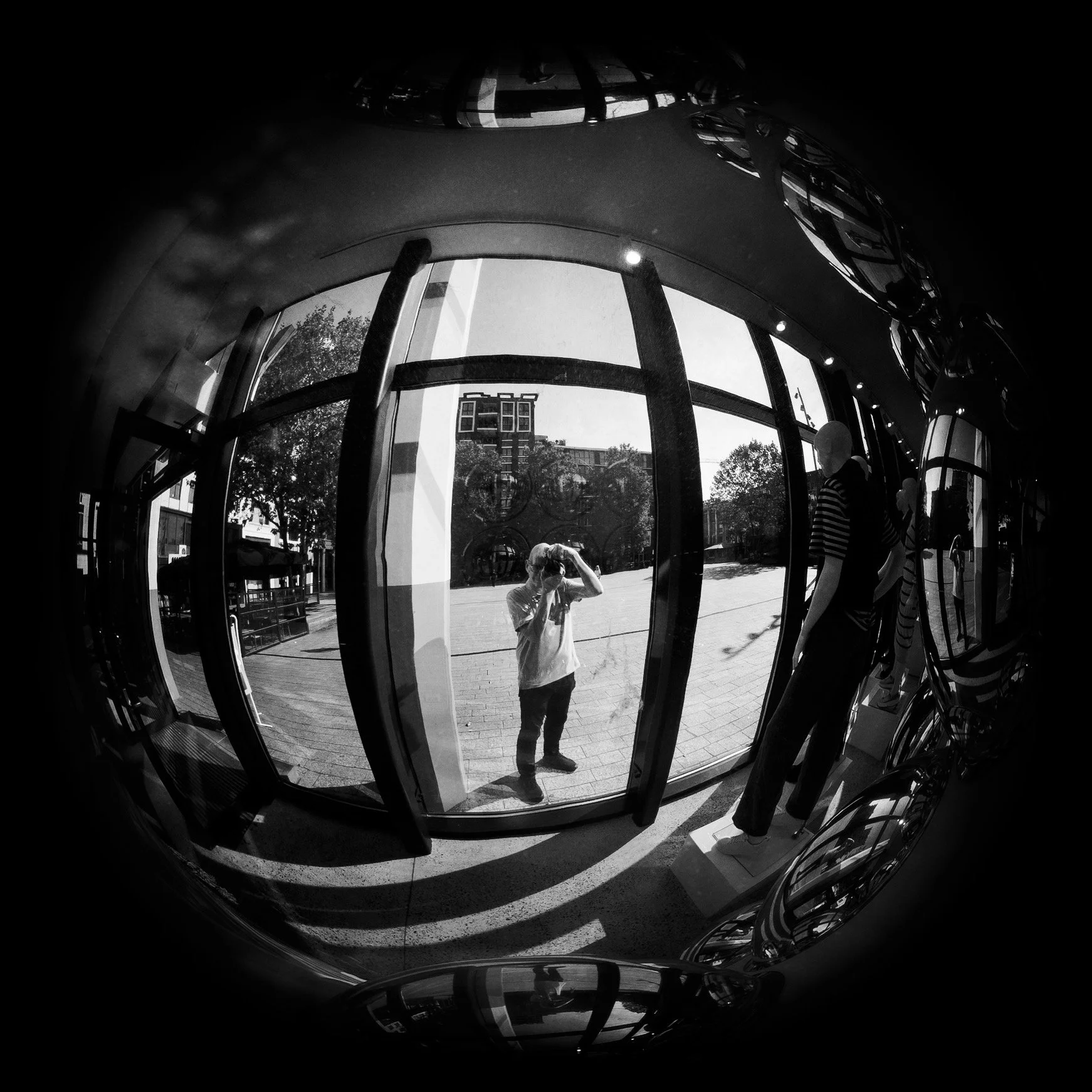 Reflection of a man taking a photo outside a large glass window, with mannequins dressed in striped clothing inside the store, city buildings, and trees visible through the window, in black and white.
