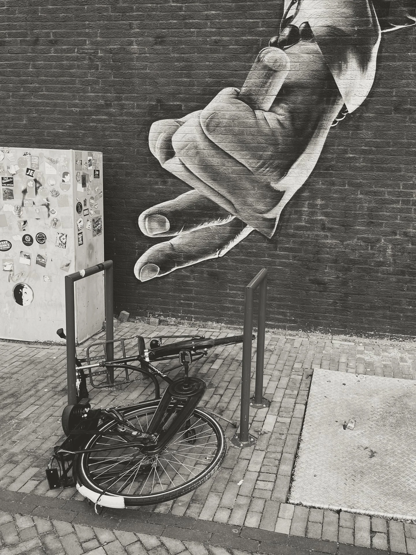 Black and white photograph of a large mural of a hand holding a smartphone painted on a brick wall. A bicycle is lying on the cobblestone sidewalk in front of the mural, with a bicycle rack nearby. There is also a small, rectangular metal platform or