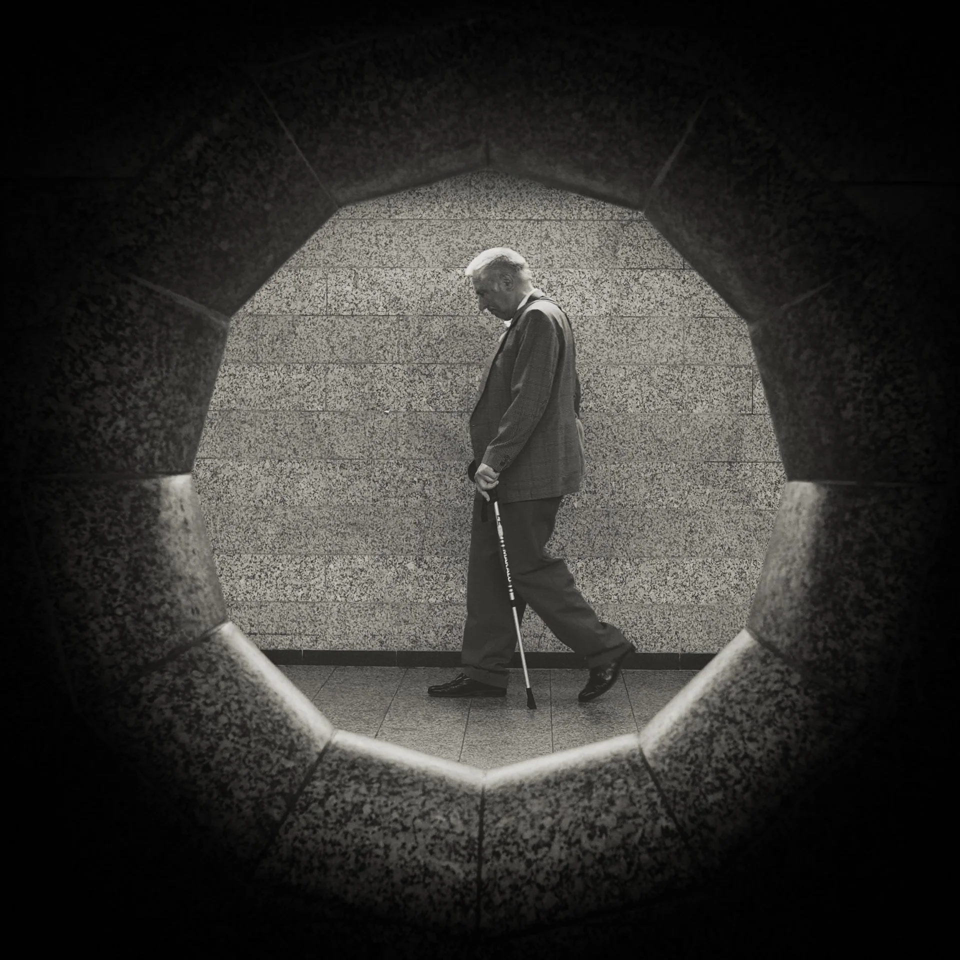 Black and white photo of an elderly man with gray hair walking using a cane, seen through a small, hexagonal opening in a stone wall.