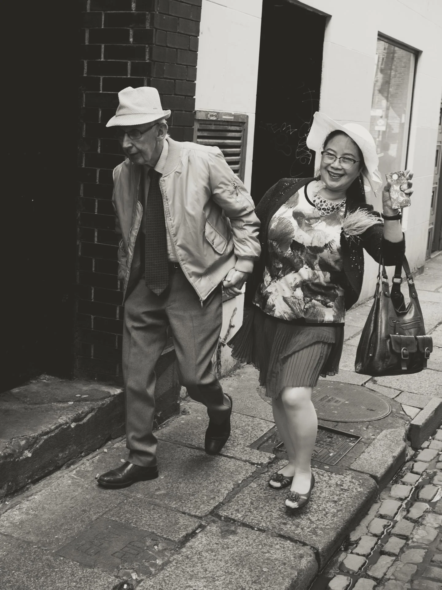 A black and white photo of an elderly man holding hands with a woman walking on a city sidewalk. The man is wearing a hat, glasses, a jacket, a shirt, trousers, and shoes. The woman is wearing a wide-brimmed hat, glasses, a colorful top, a skirt, sto
