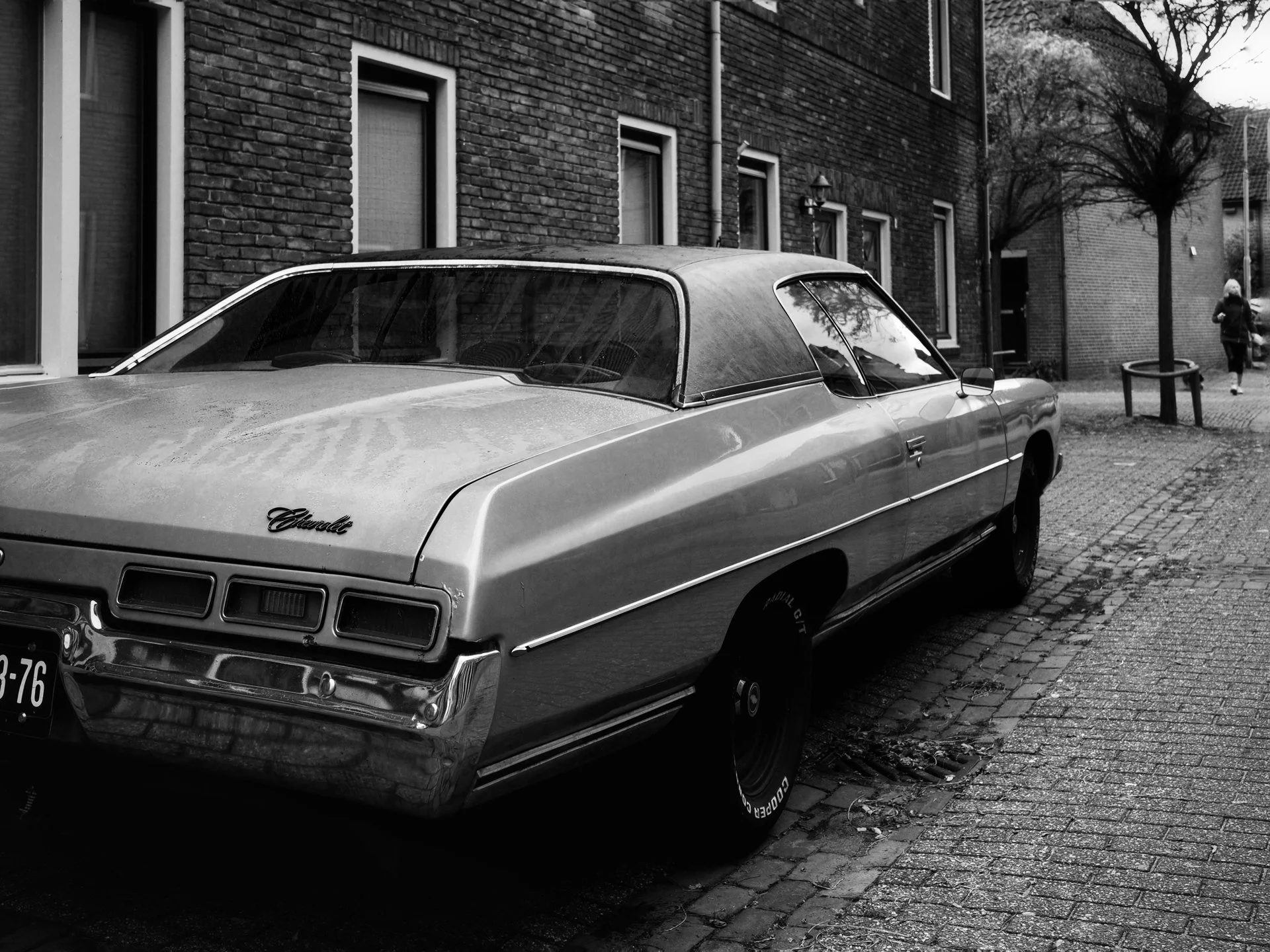 A vintage Chevrolet car parked on a cobblestone street next to brick buildings, with a person walking in the background.