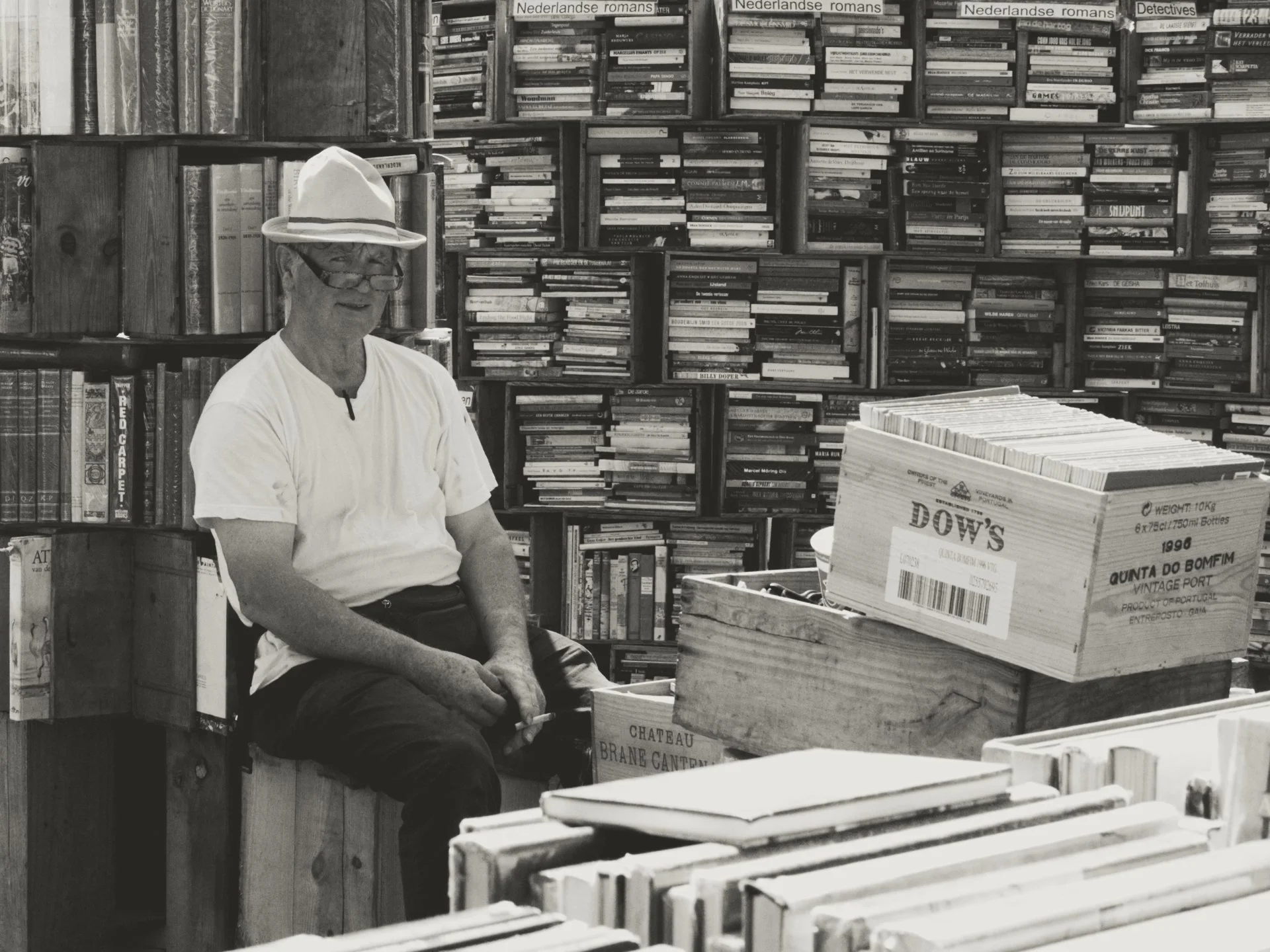 An elderly man wearing a white hat, glasses, and a white shirt sitting in a bookstore surrounded by shelves filled with books. There are boxes of books in front of him.