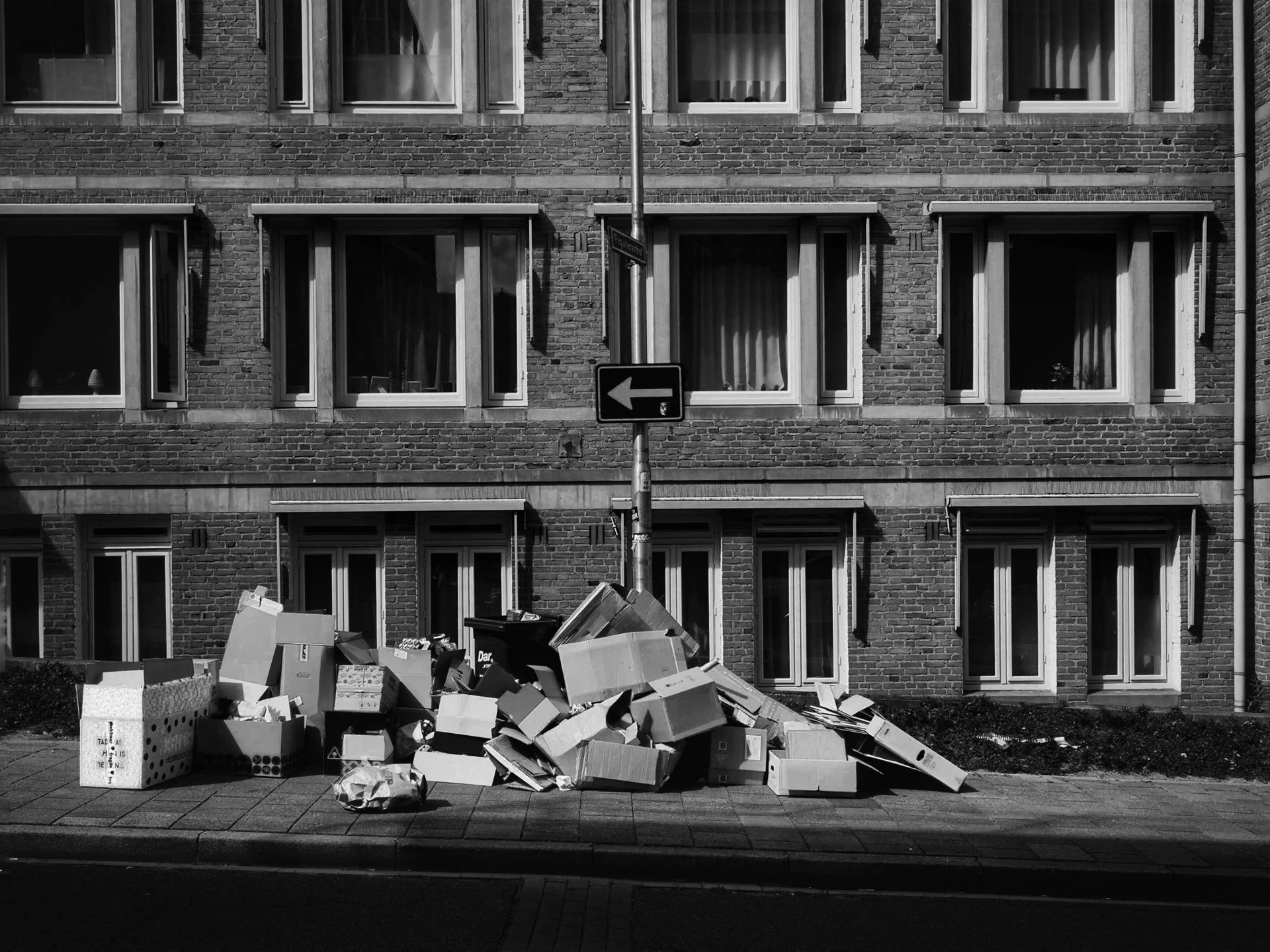 A pile of cardboard boxes and trash on the sidewalk in front of a brick apartment building with multiple windows.