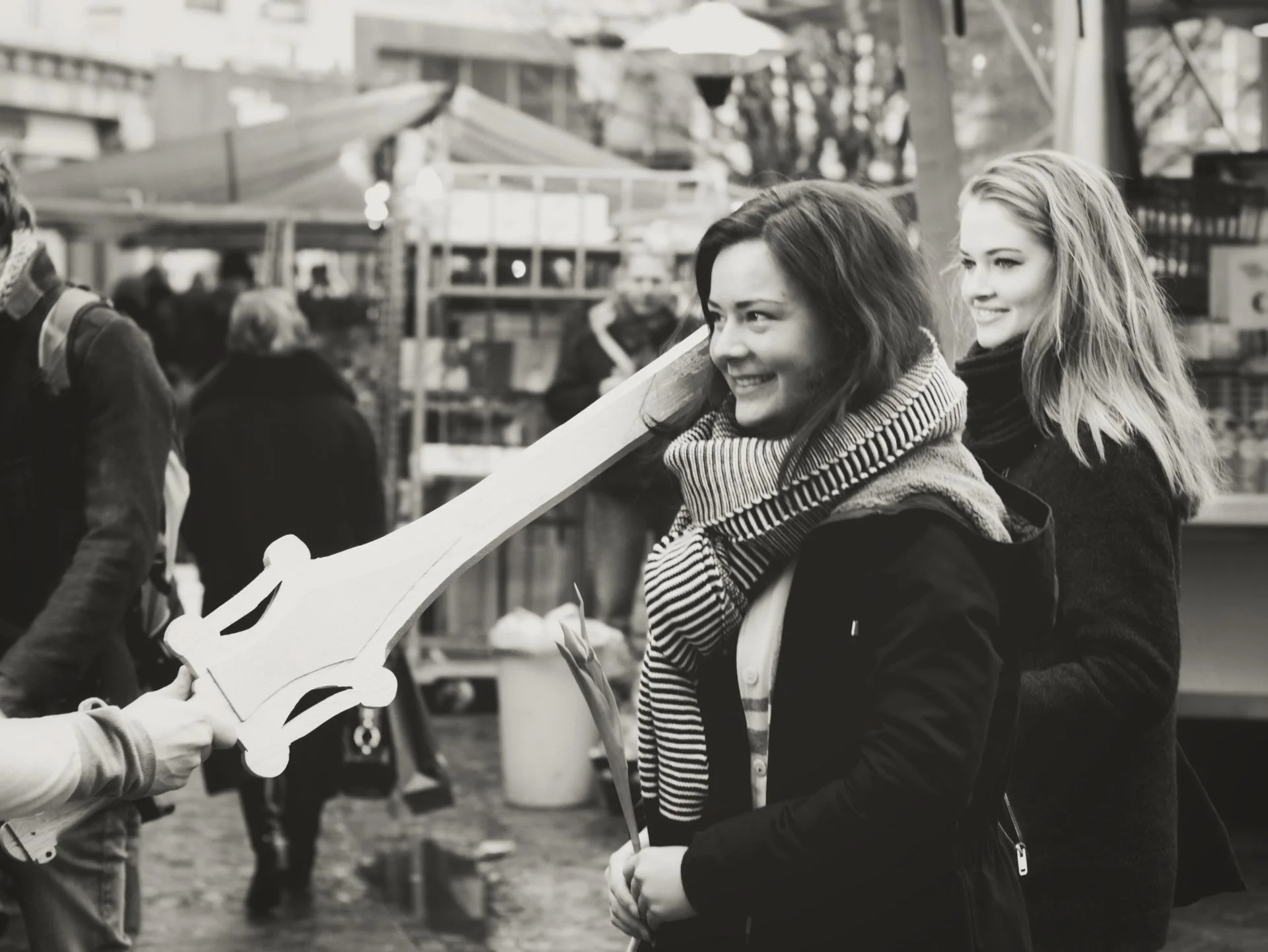 Two women smiling at each other at an outdoor fair or market, with one woman holding a paper craft and another person handing her something.