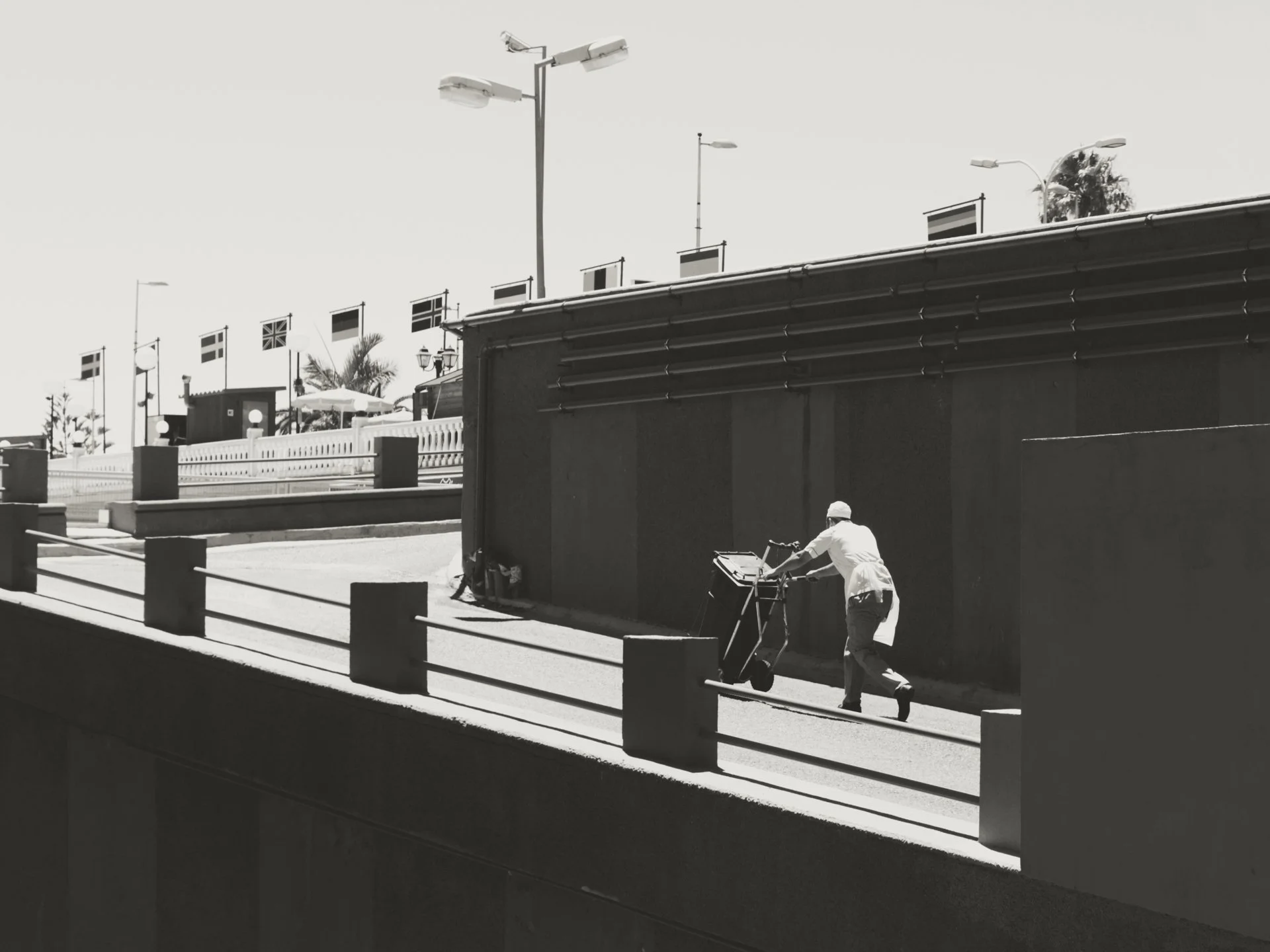 A person wearing a white jacket and hat pushes a hand truck with a basket along a sidewalk, with a modern building and flagpoles in the background.