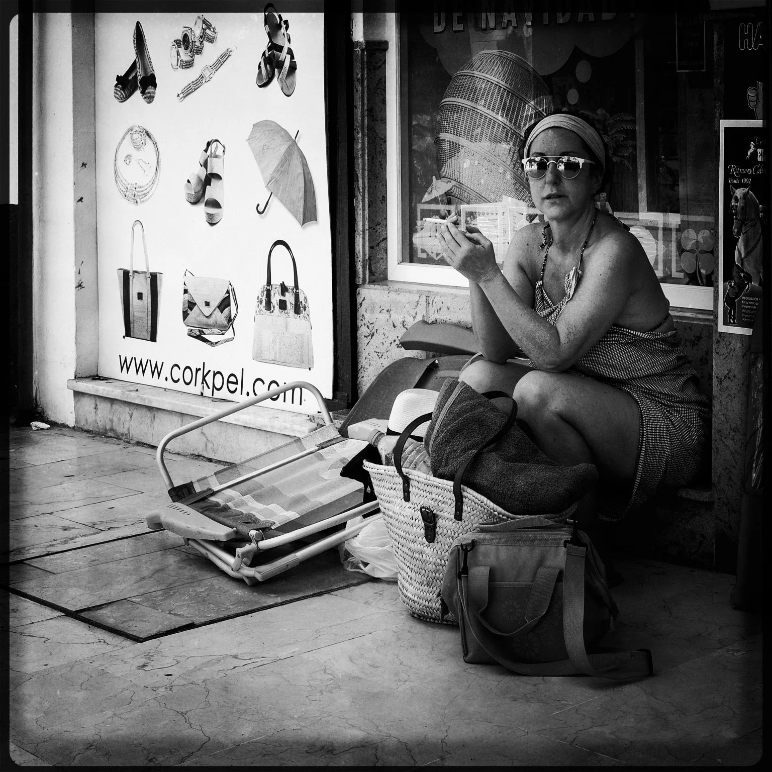 A woman with sunglasses and headscarf sitting on a bench beside shopping bags, a foldable chair, and a large tote bag, outside a store with fashion advertisements.