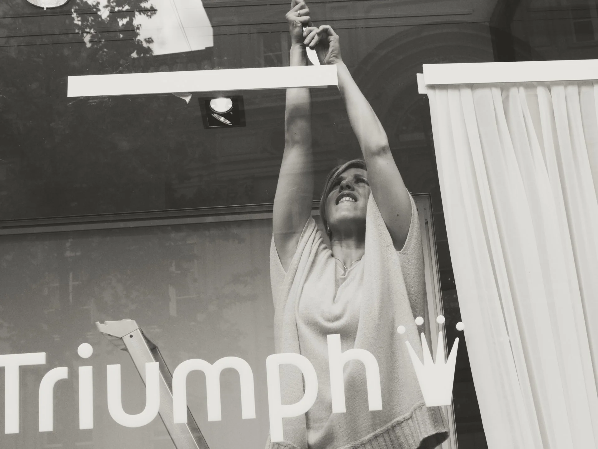 A woman cleaning glass with a cloth, seen through a window with some text and reflections of trees and a building in the background.