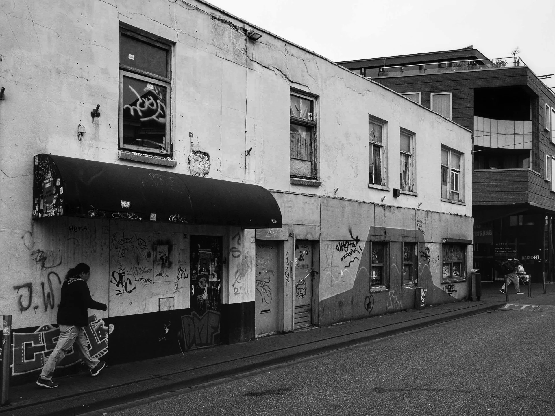 A black and white photo of a graffiti-covered building with a person walking with a backpack on the sidewalk.