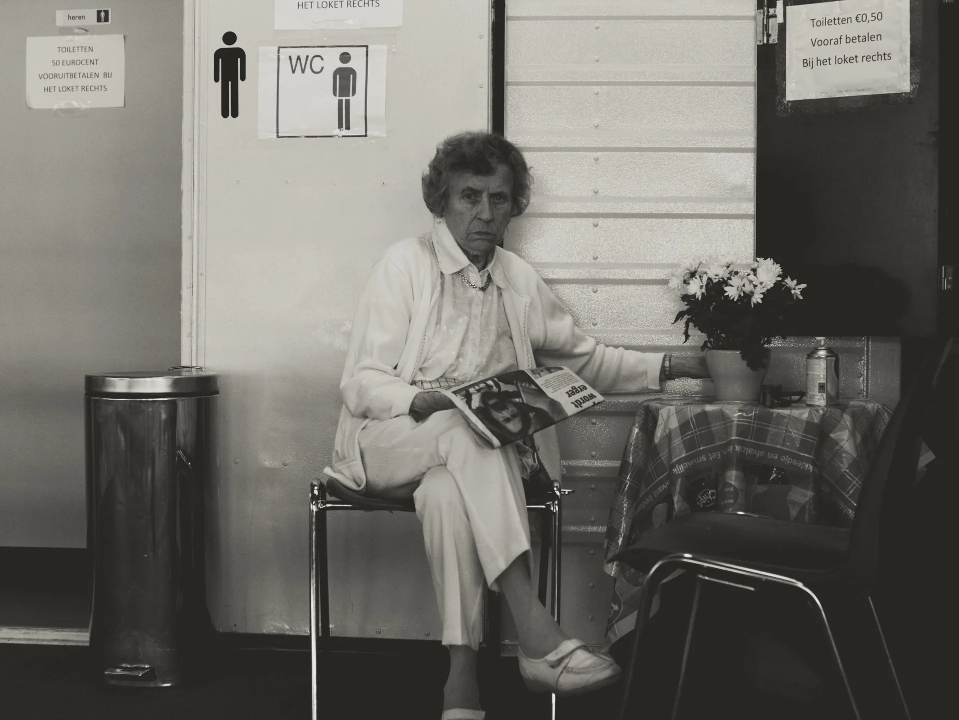 An elderly woman sitting in a wheelchair at a table in a public space, holding a magazine. She is dressed in a white outfit with a cardigan and is wearing white shoes. There are signs in Dutch on the wall, a small table with a checkered cloth, a pott