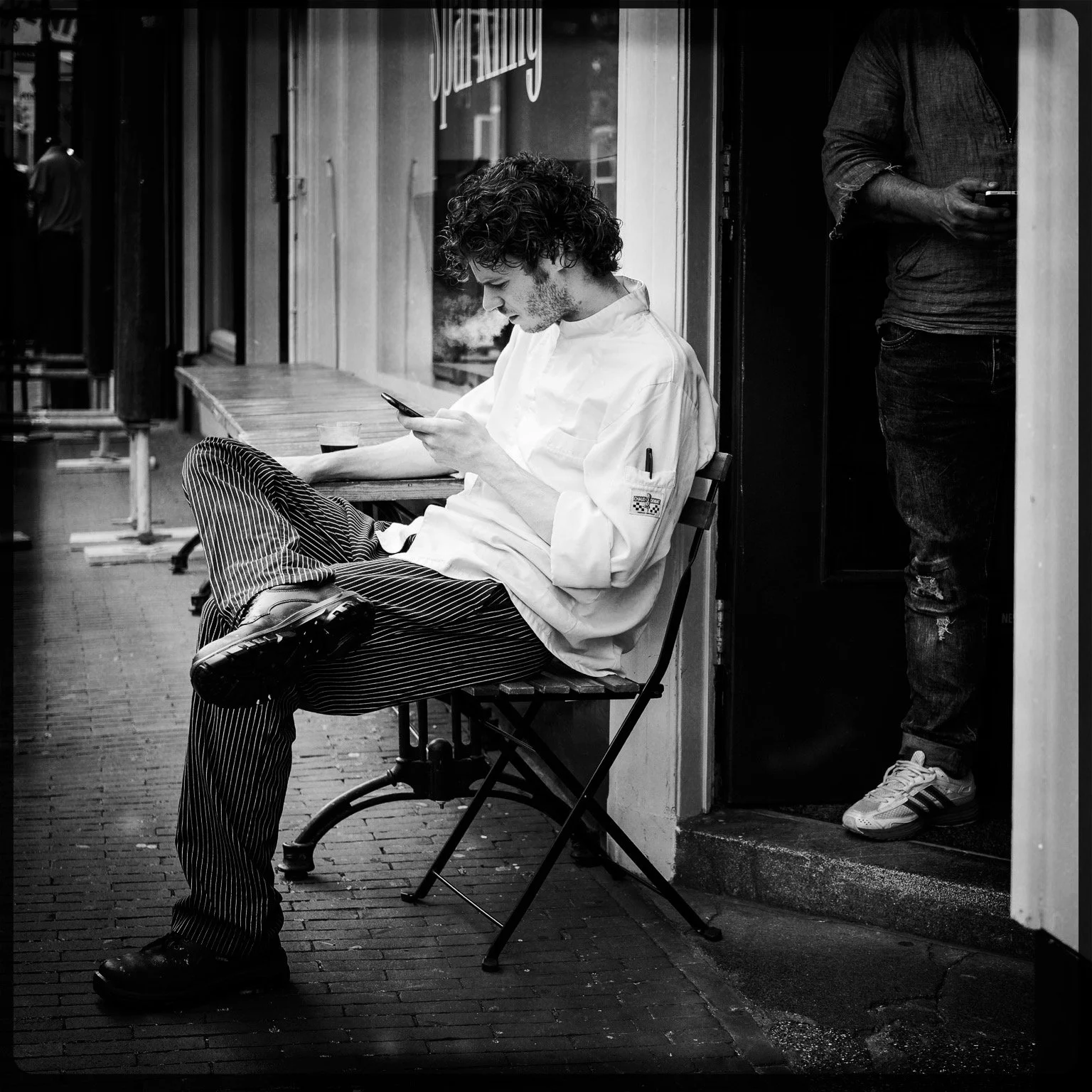 A young man with curly hair, wearing a white chef's coat and striped pants, sitting on a folding chair outside a cafe, looking at his phone. Another person is partially visible standing inside the doorway, also using a phone.