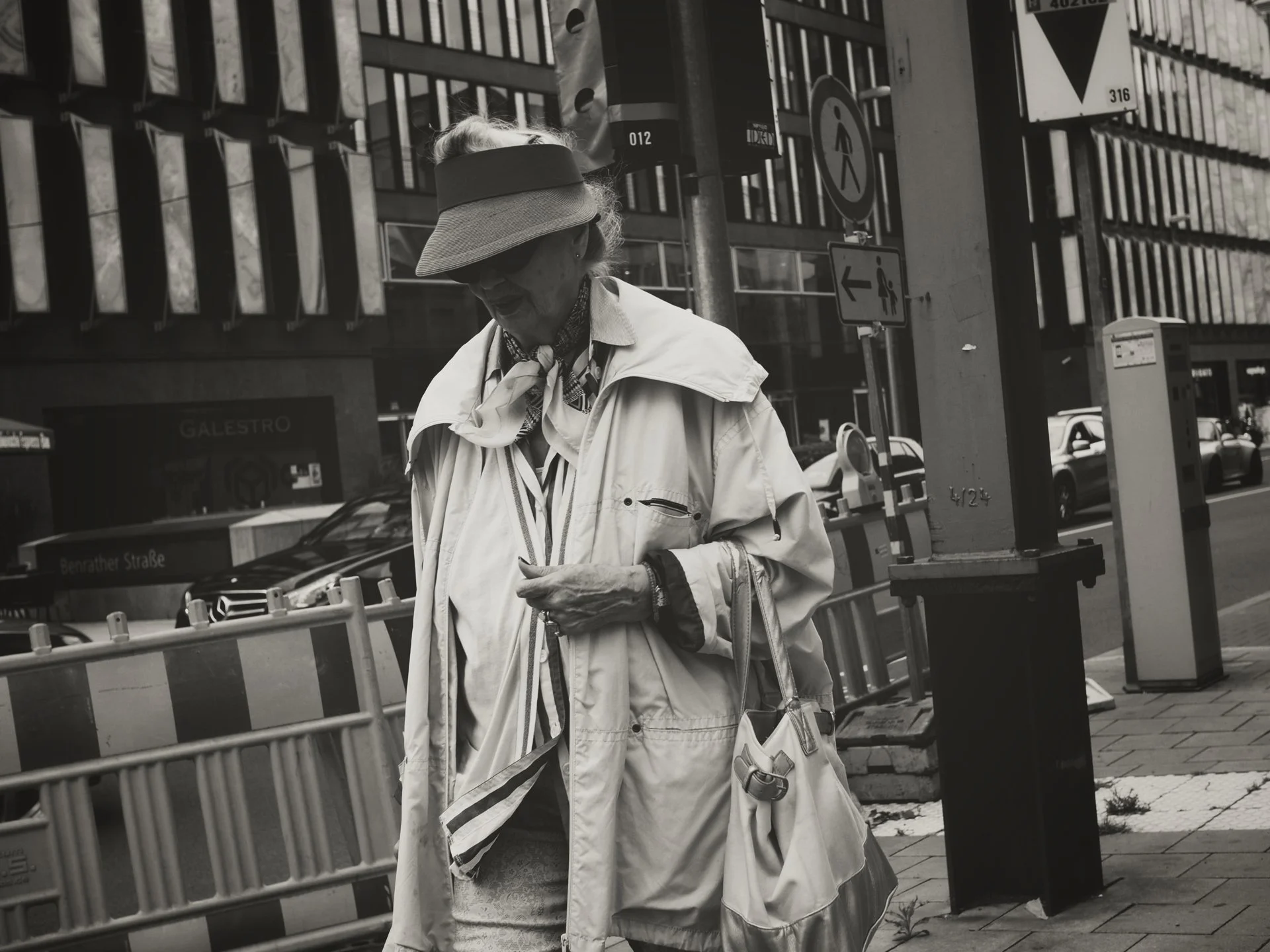 An elderly woman wearing a visor and light-colored jacket stands on a city sidewalk, looking down at her phone. She is surrounded by street signs, cars, and a building with modern architecture in the background.