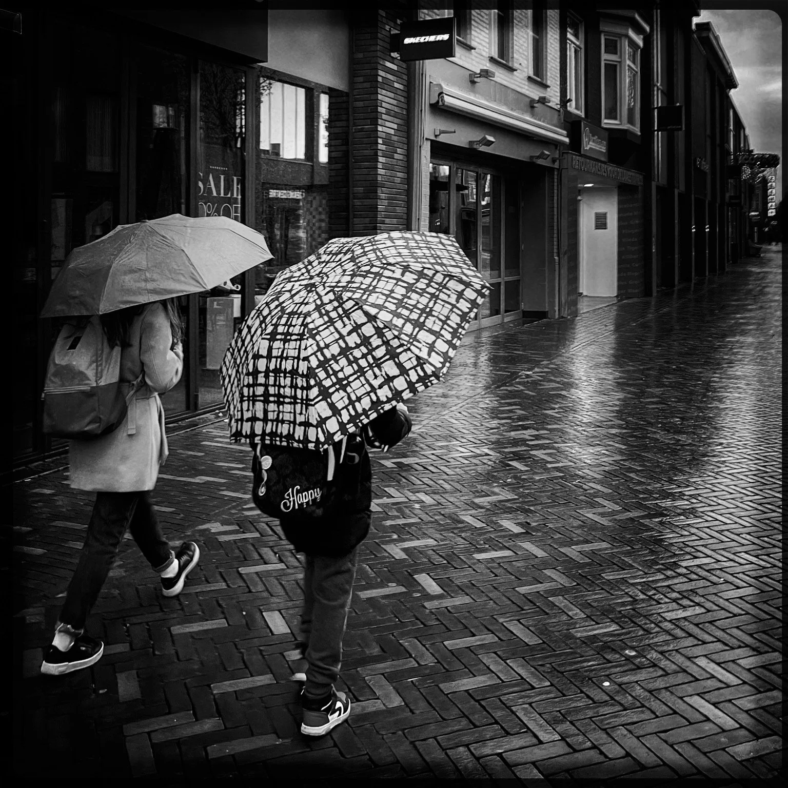 Two children walking on a wet city sidewalk with umbrellas during a rainstorm, storefronts and buildings in the background.