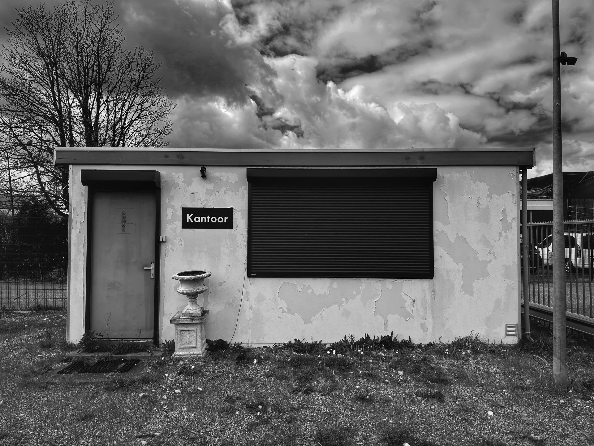 A small, weathered building with a closed shutter window, a door, a sign that reads 'Kantoor', a birdbath, and a leafless tree in the background under a cloudy sky.