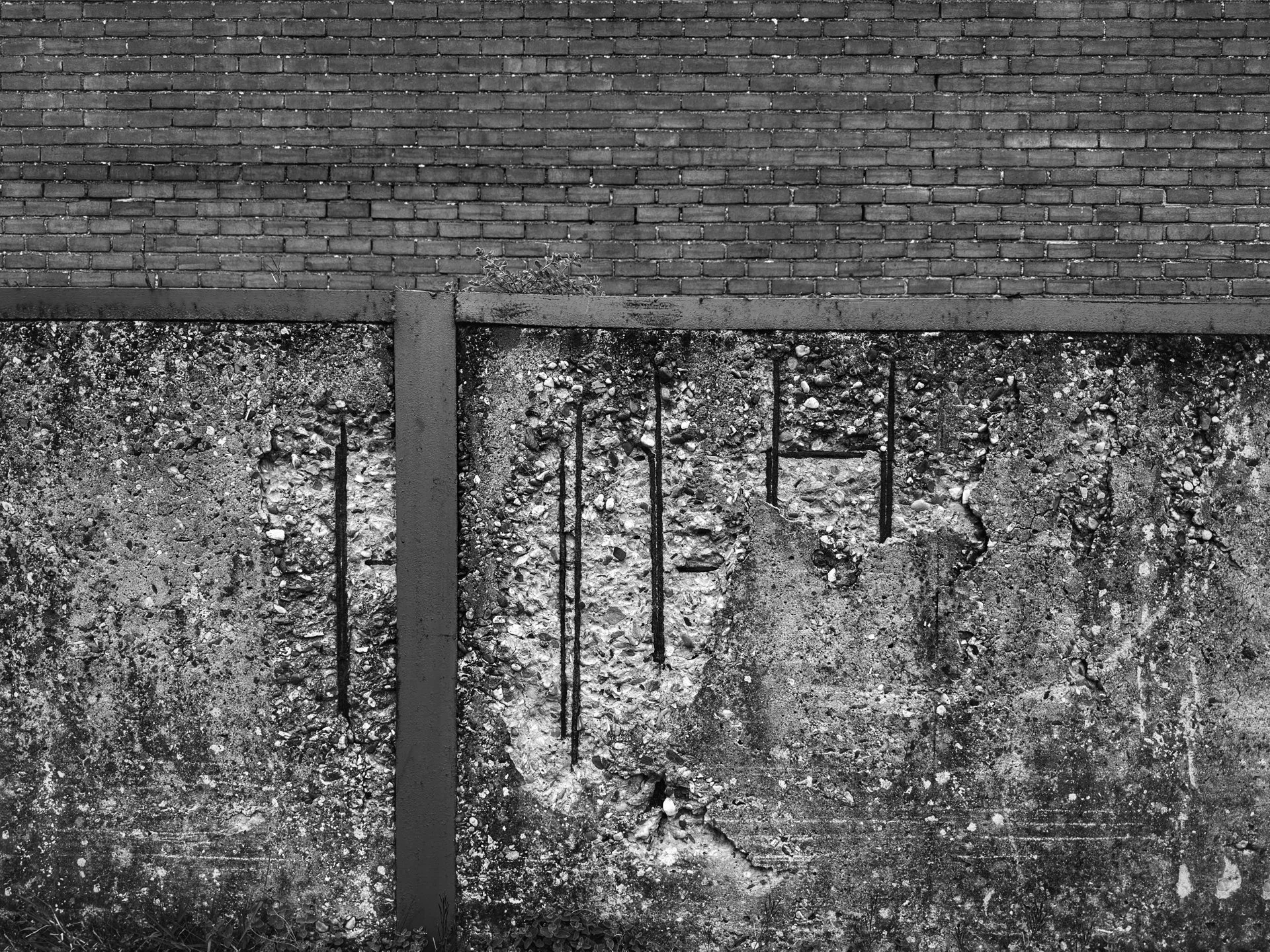 A black and white photo of a brick wall with a separated section of a weathered concrete wall below it, which has rusted metal reinforcement bars visible.