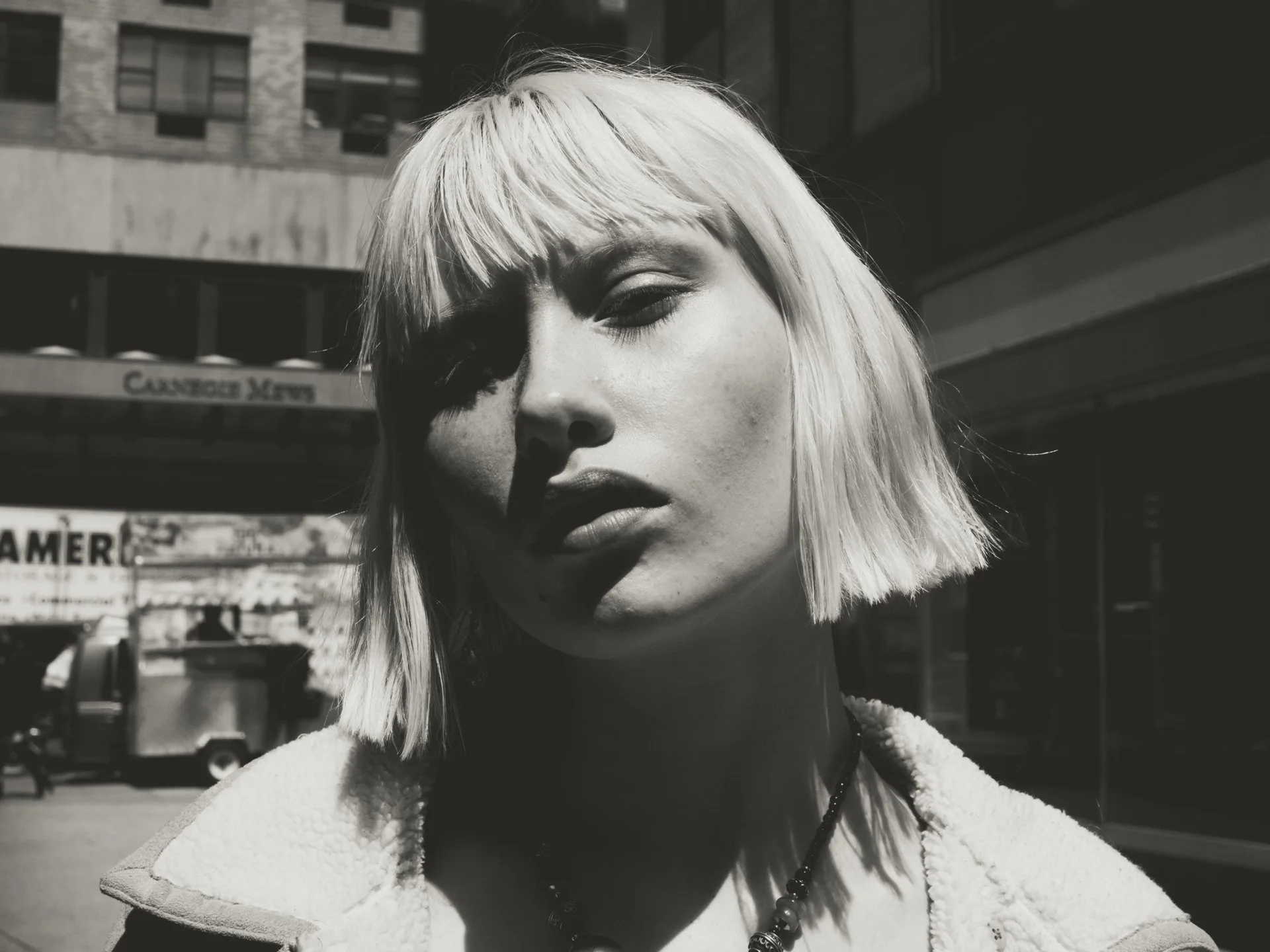 A black-and-white close-up portrait of a woman with platinum blonde hair, wearing a fluffy jacket and a beaded necklace, standing outdoors with urban buildings and a sign that says 'Cambridge News' in the background.