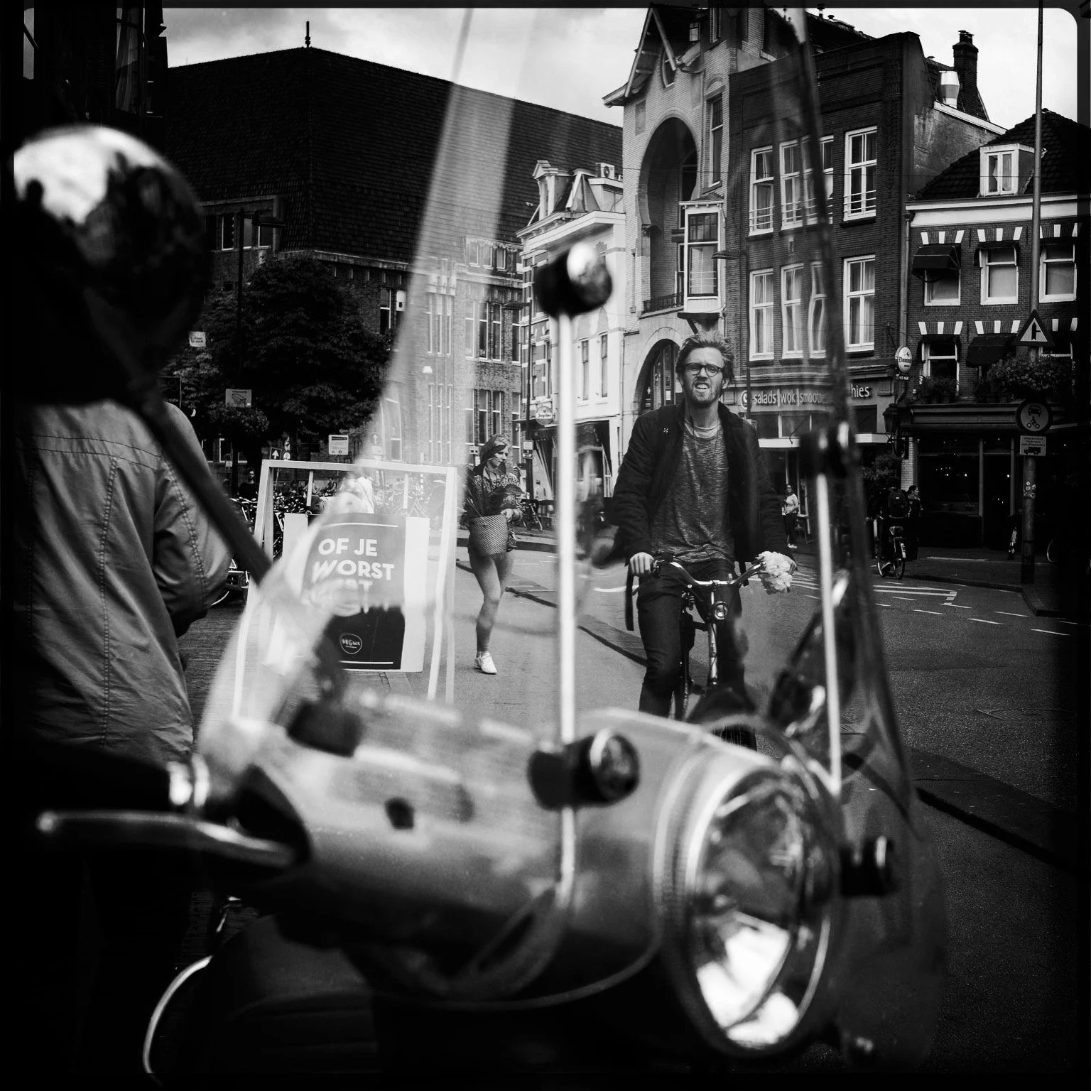 Black and white street scene with a man riding a bicycle and smiling, seen through a parked motorcycle's windshield. Pedestrians walking along the sidewalk, buildings with varied architectural styles, and street signs are visible.