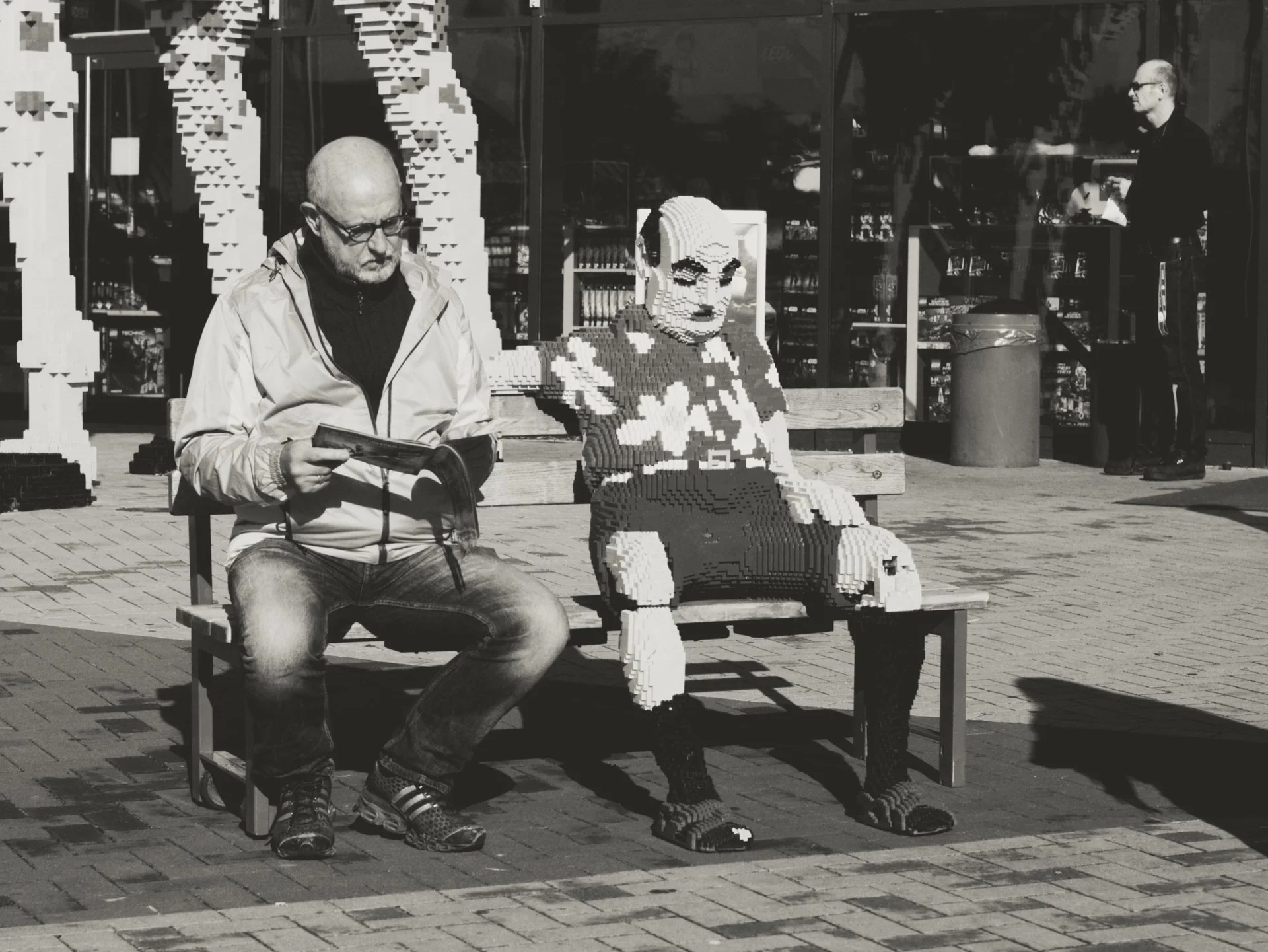 A man with glasses and a bald head wearing a light-colored jacket and jeans is sitting on a bench, reading a magazine. Next to him is a life-sized sculpture made of LEGO bricks that depicts a woman in sunglasses, a patterned shirt, and pants, seated 