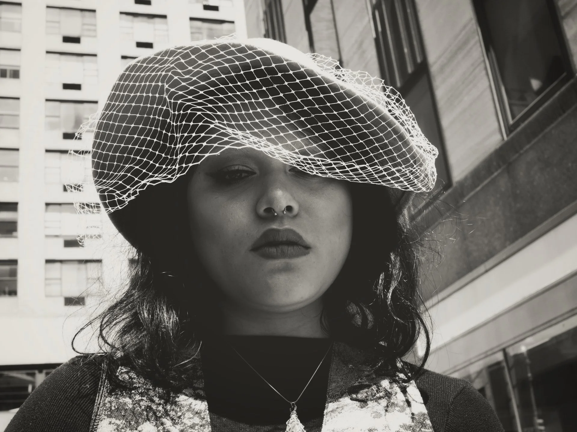 A woman with dark wavy hair wearing a netted hat and lipstick, standing outdoors in front of a building with windows.