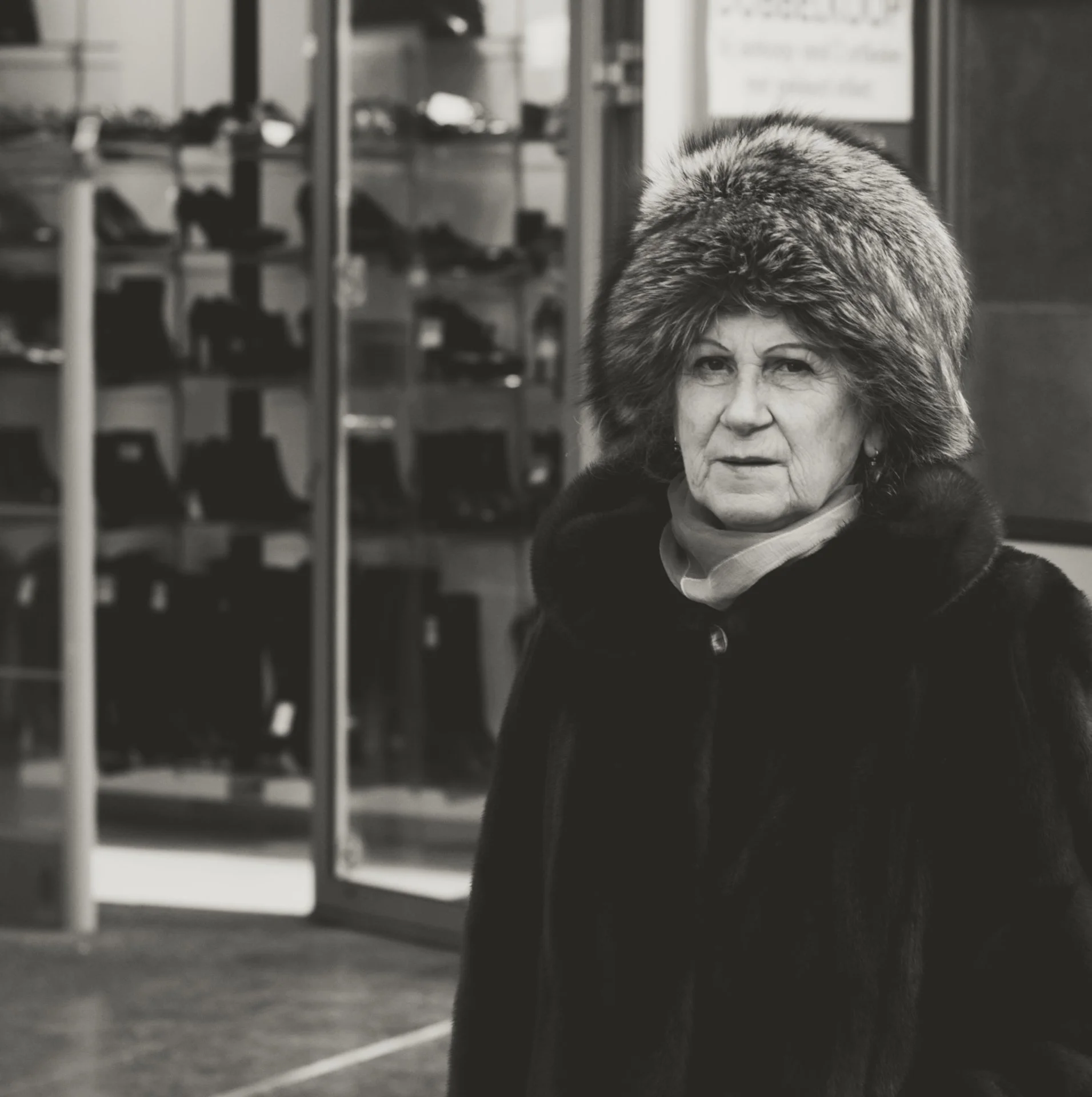 Black and white photo of an older woman with a large fur hat, sitting indoors with shelves and furniture in the background.