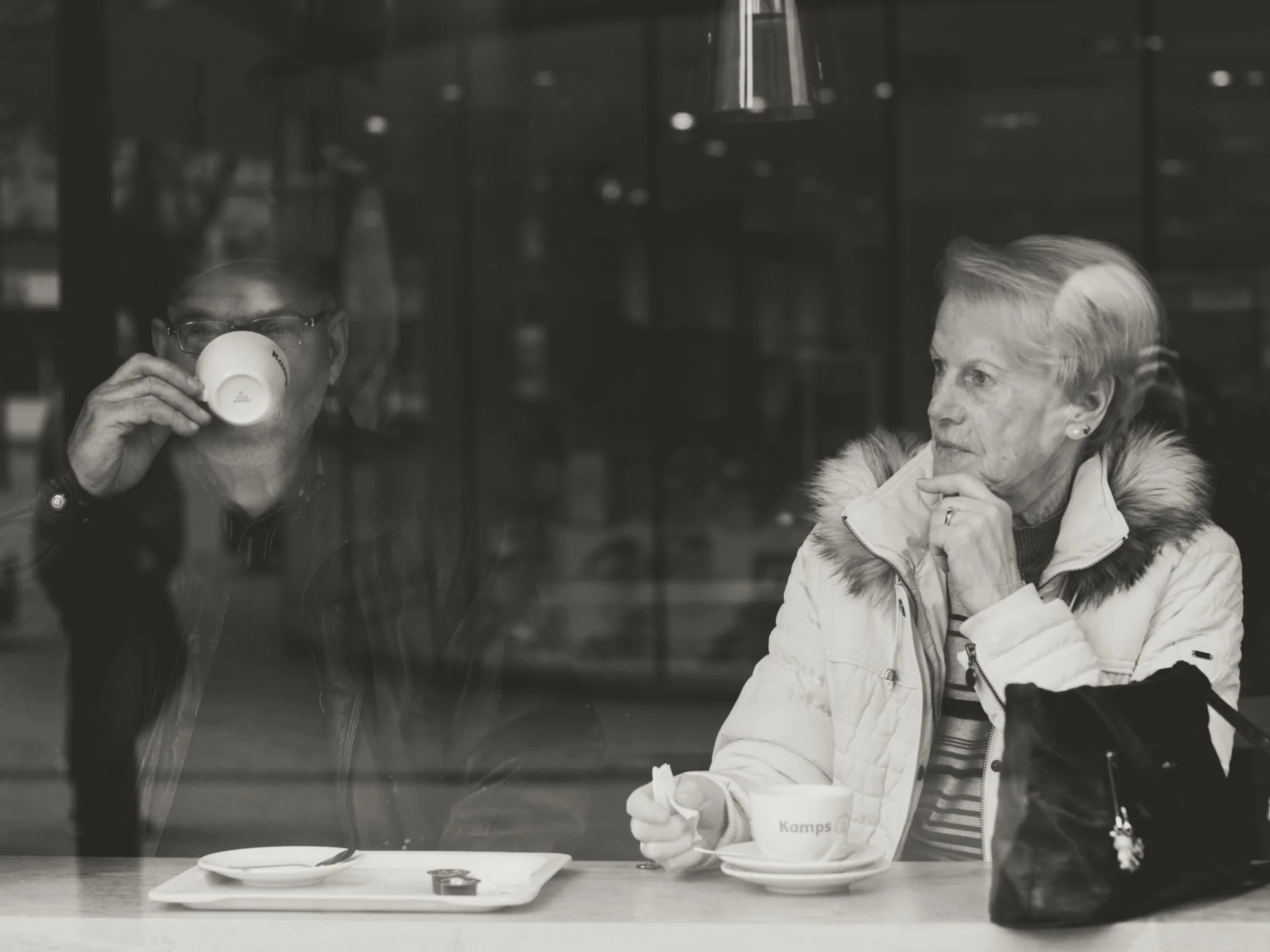 A man with glasses drinking from a white mug and a woman in a white jacket sitting at a table with a cup of coffee or tea, seen through a glass window in a cozy cafe.