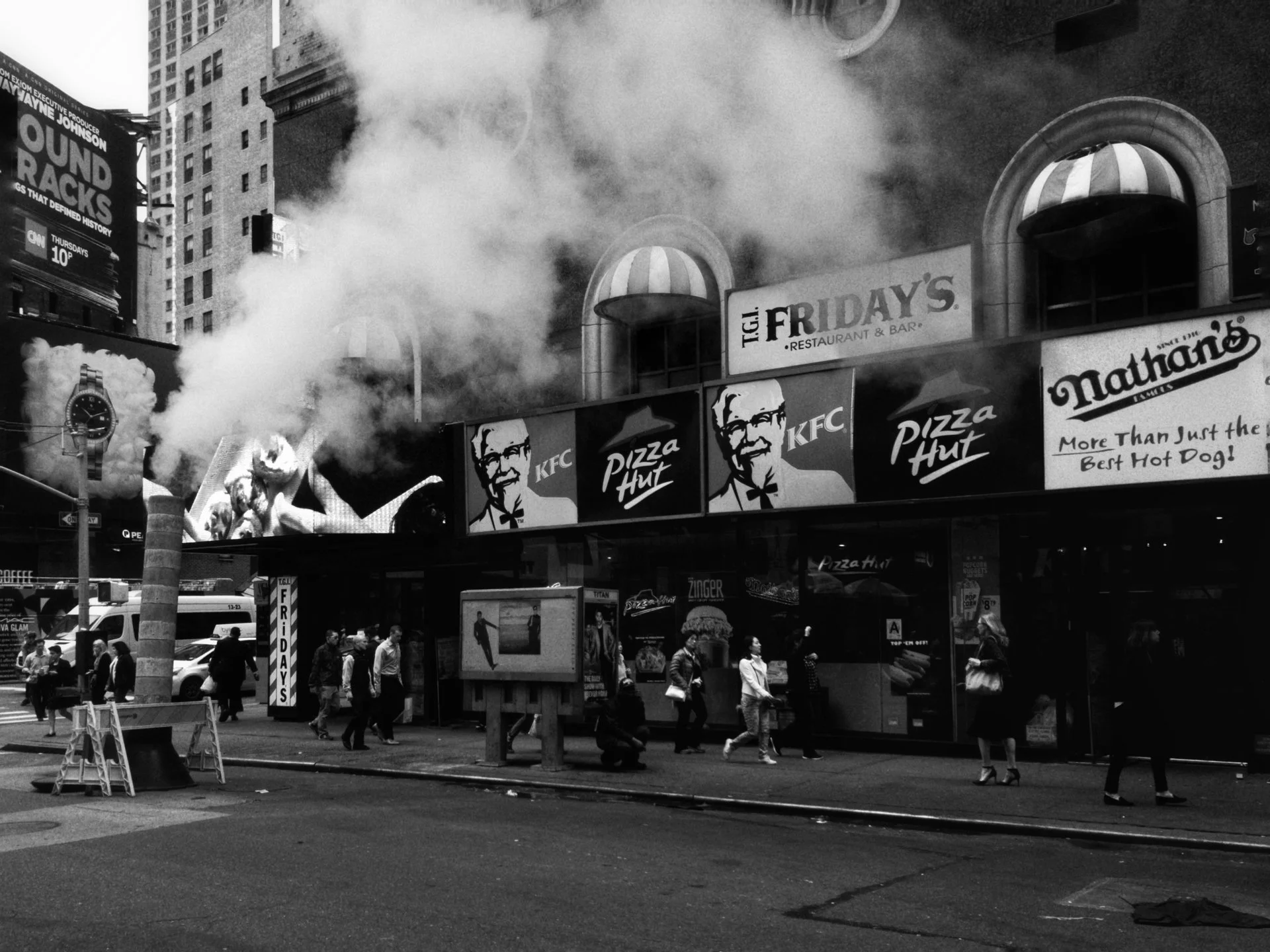 A city street scene with people walking in front of a fast food restaurant. Smoke is billowing from a chimney on the building, and there is a digital billboard displaying a watch advertisement, along with a sign for T.G.I. Friday's restaurant. The sc