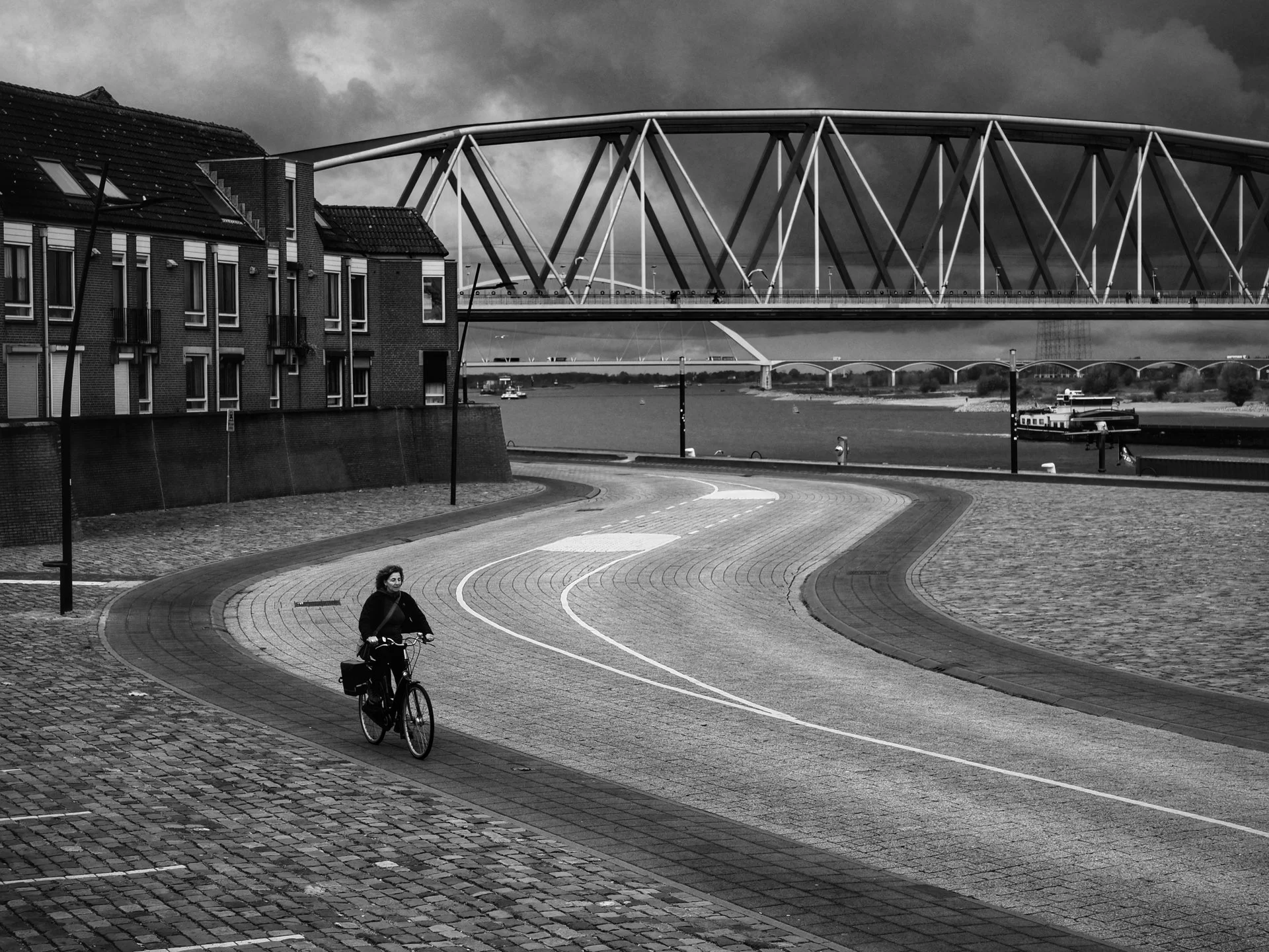 A woman biking on a curving paved road near a river with a bridge overhead and buildings on the side, under a cloudy sky.