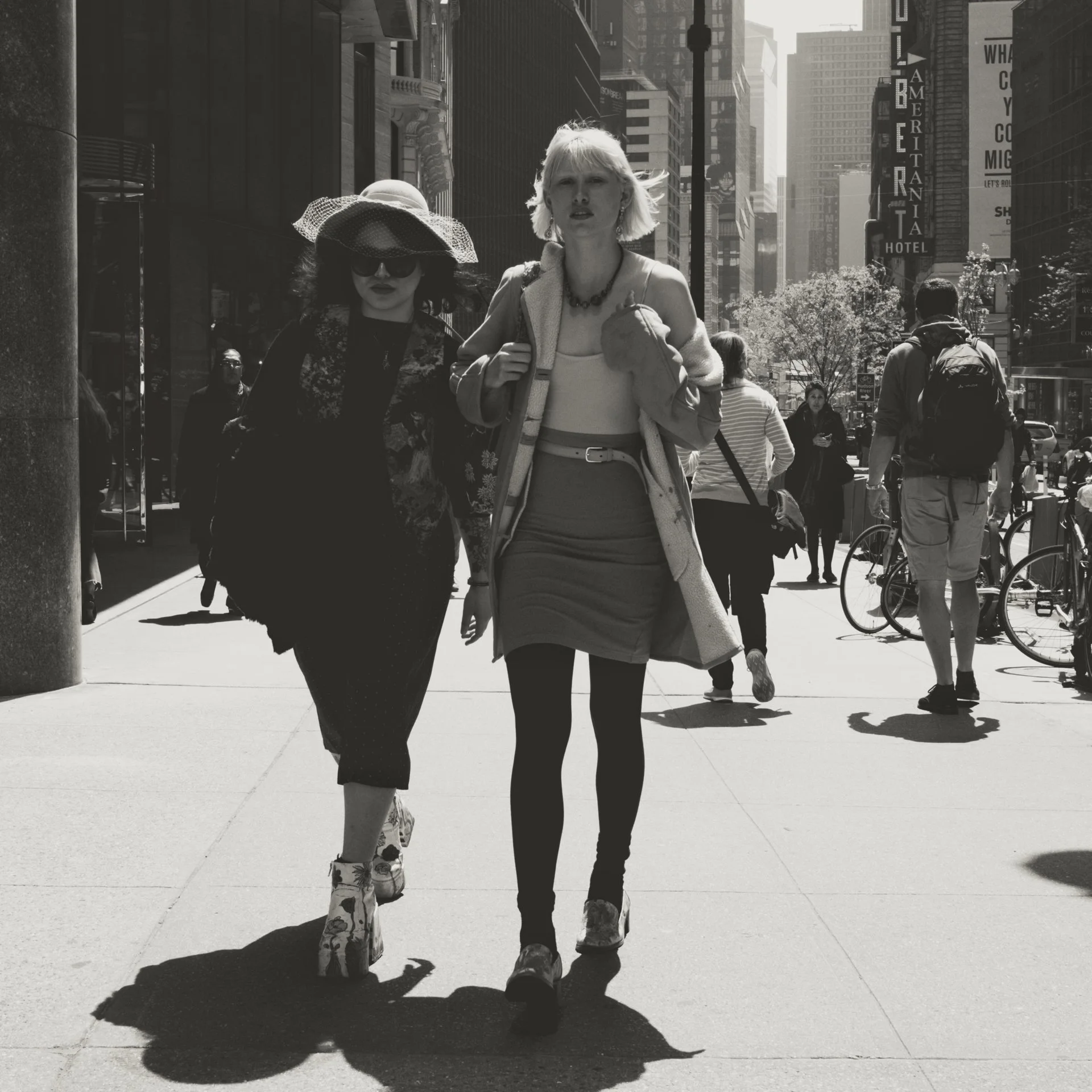 Two women walking in a city street during daytime, with tall buildings and people in the background. One woman wears a large sun hat and sunglasses, the other has short blonde hair and is dressed in light clothing.