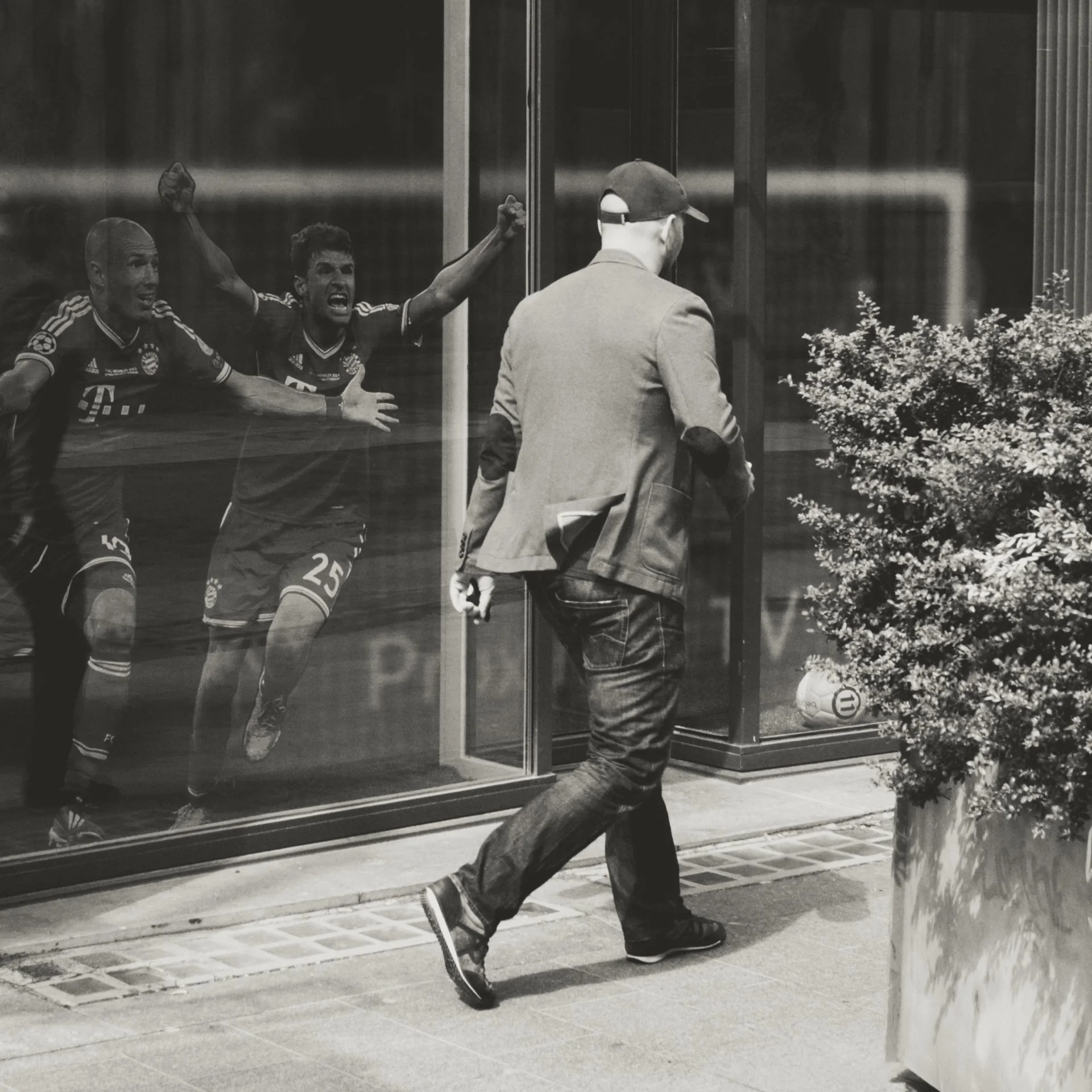 A man in a blazer and jeans walking past a glass window with a reflection of two soccer players in Bayern Munich jerseys celebrating, with one player raising his arm and the other showing a thumb up. There are plants outside and a sidewalk in front o