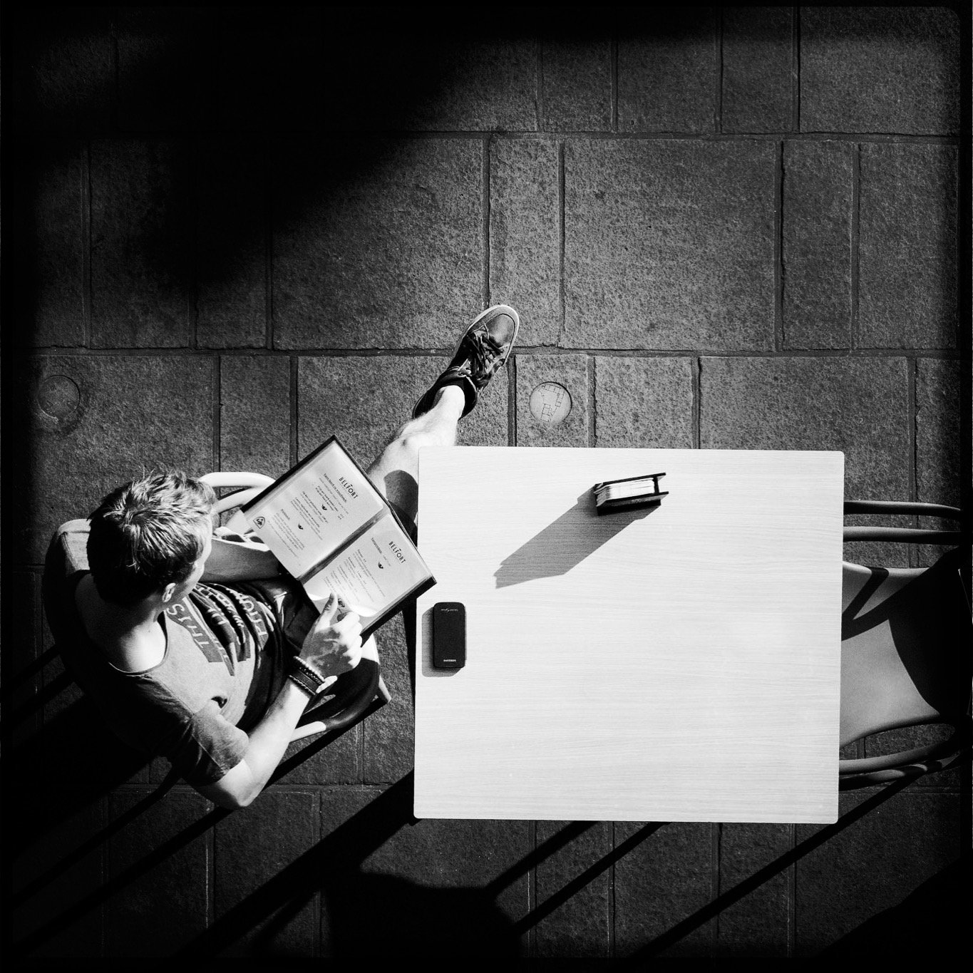 A person sitting at a table reading a menu with a smartphone and a book on the table, seen from above. The scene is in black and white.