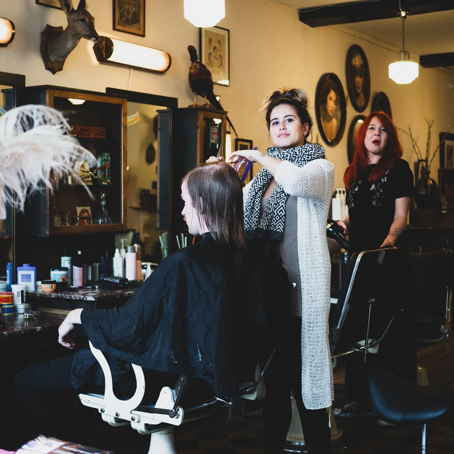 A hairdresser is cutting a woman's hair in a vintage-style hair salon with framed photographs and mounted animals on the wall.