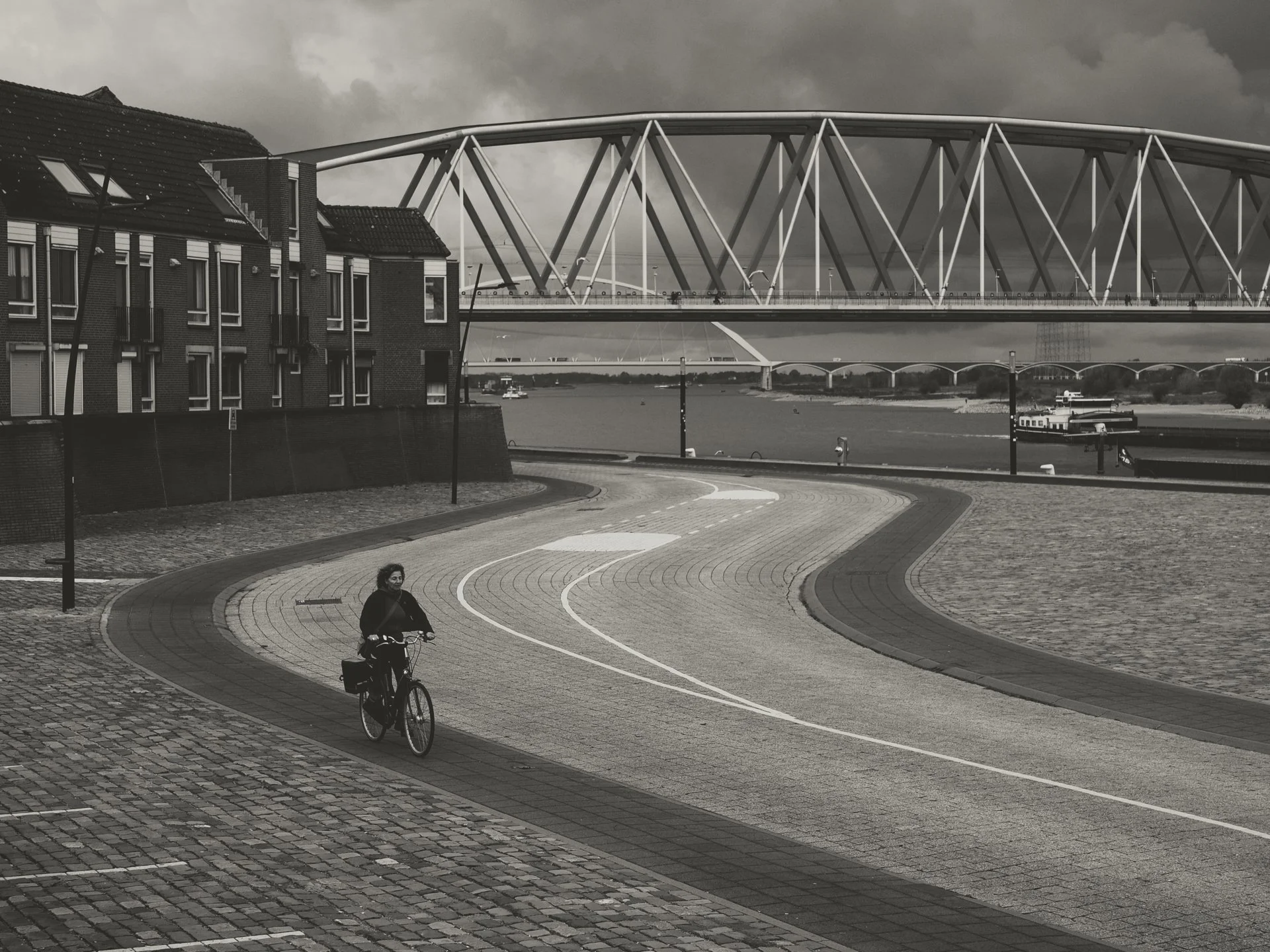 A black and white photo of a woman riding a bicycle on a paved trail near a body of water. In the background, there is a bridge with a modern, geometric design and several boats on the water. To the left, there are residential buildings with sloped r