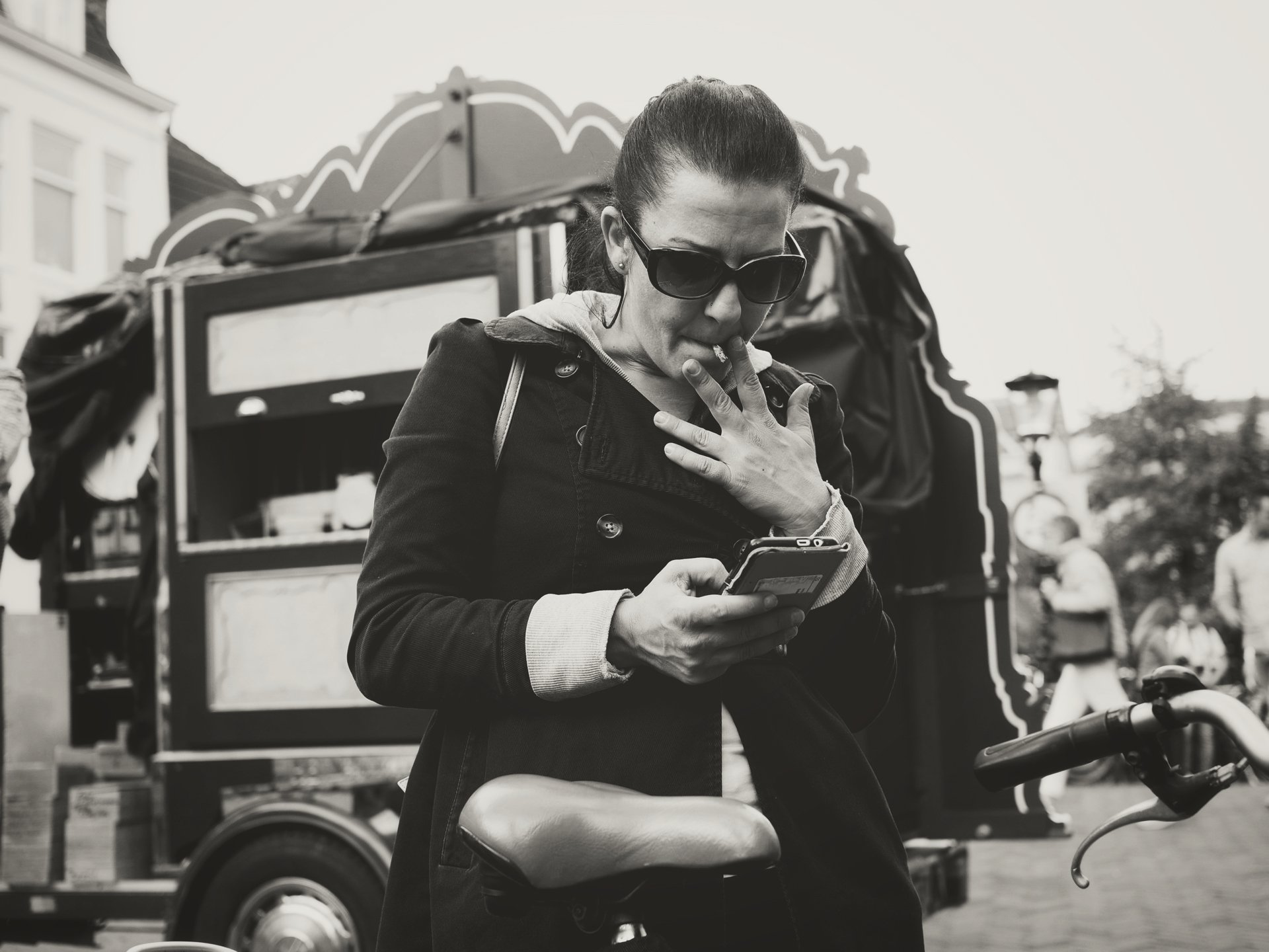 A woman wearing sunglasses and a dark jacket is looking at her phone while smoking a cigarette. She is outdoors, with a food cart and people in the background.