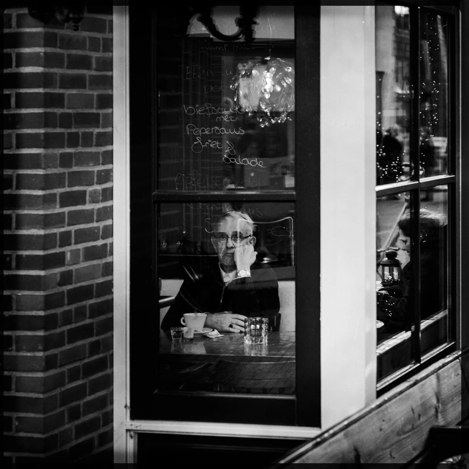 A black and white photo taken inside a cafe or restaurant, showing a man sitting at a table, viewed through a window. Inside, he appears to be resting his head on his hand, with a coffee cup and water glass on the table. The window has handwritten me
