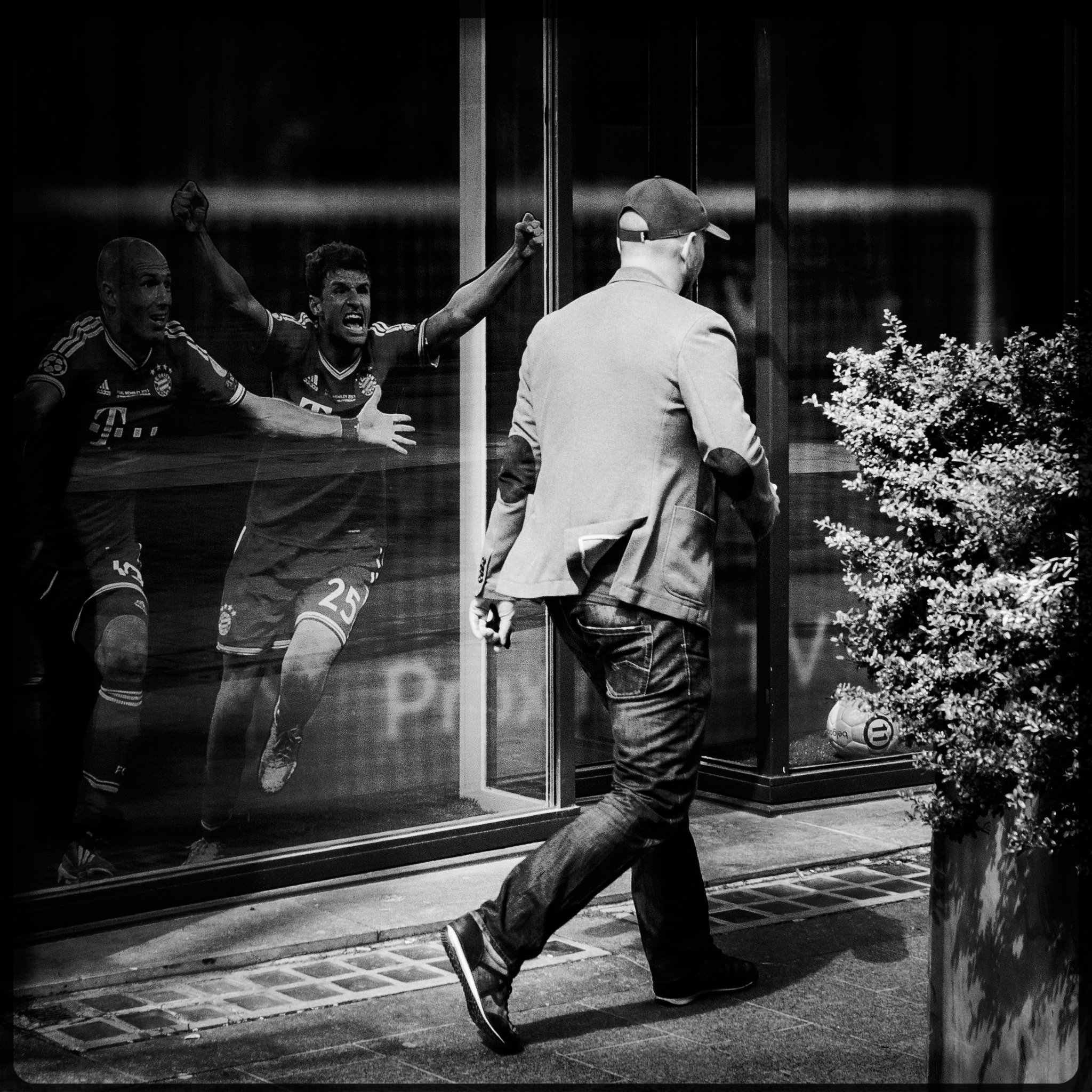 A man in a baseball cap and jacket walking past a glass window with soccer players' images inside. The players are celebrating, and the scene is outdoors with a plant nearby.