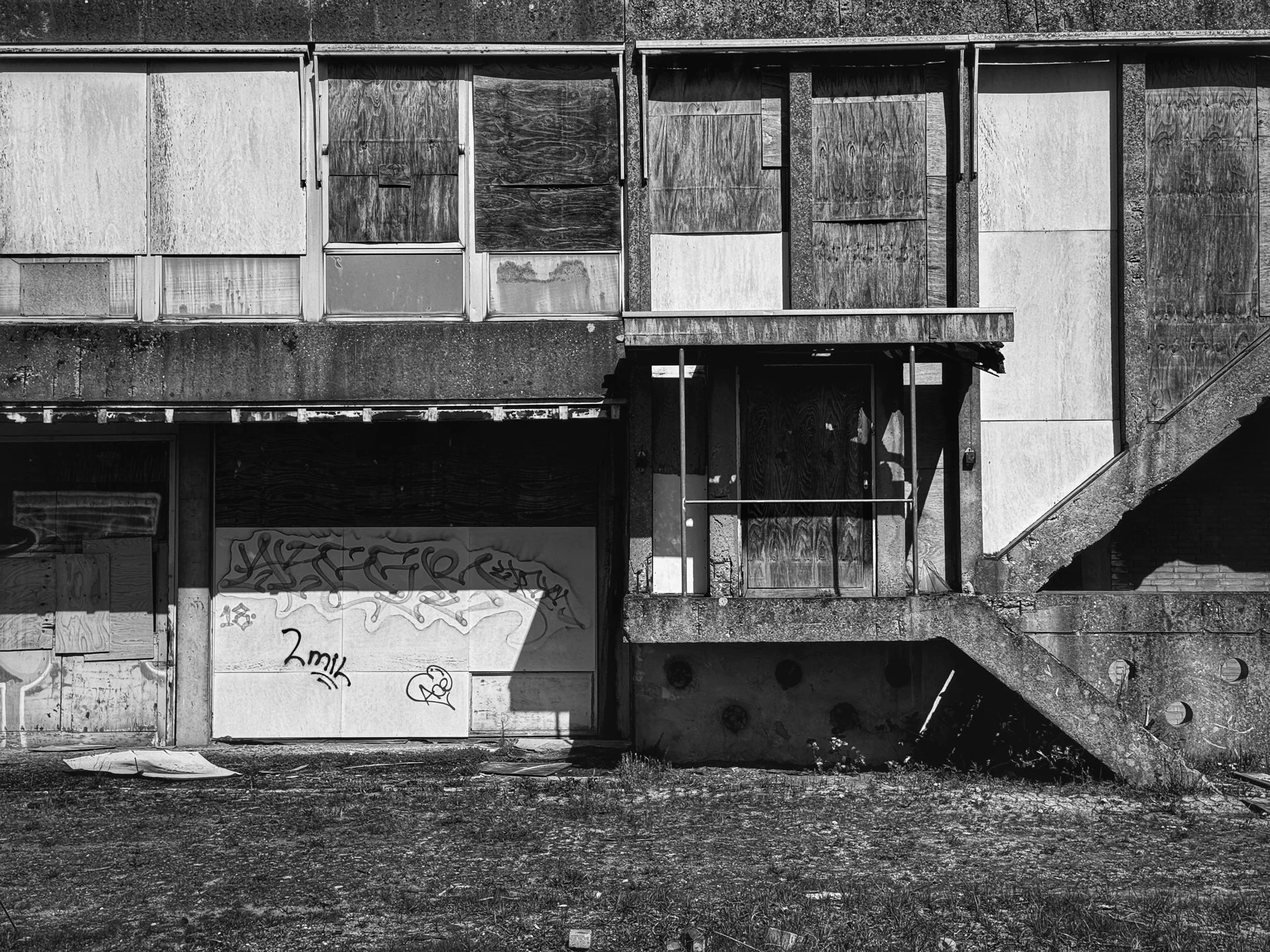 An abandoned building with boarded-up windows and a staircase, graffiti on the lower wall, and a patch of grass and dirt in front.