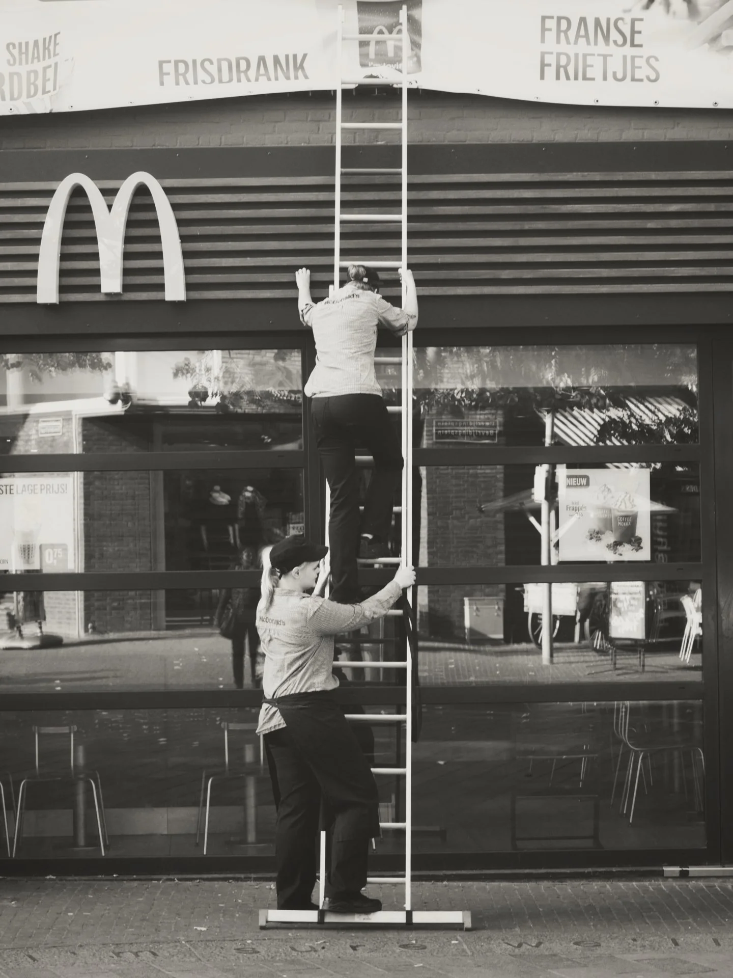 Two women in uniform are cleaning windows from outside a McDonald's restaurant, one standing on the ground holding a ladder, and the other climbing the ladder with cleaning tools.