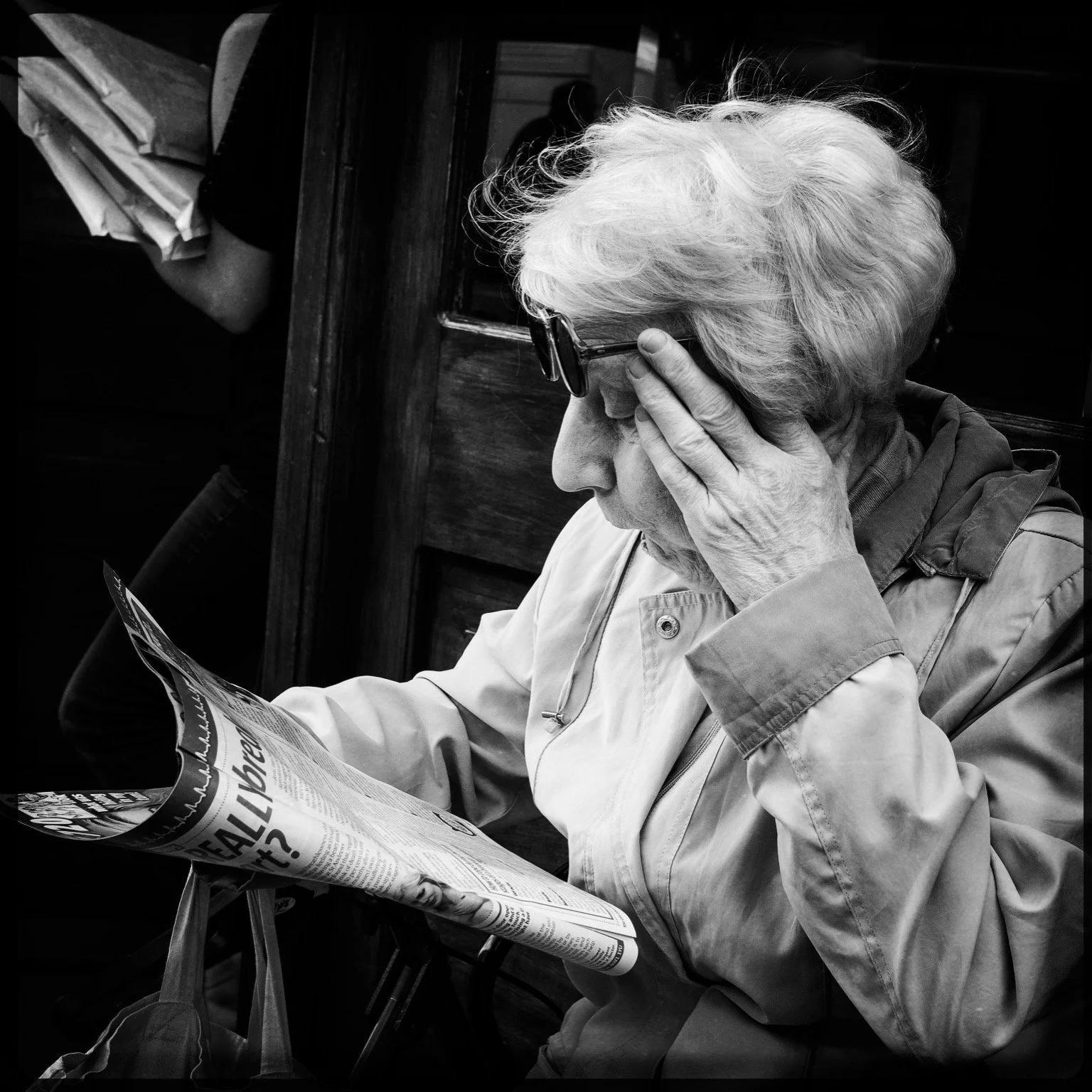 An elderly woman with glasses is reading a newspaper, holding her forehead with her left hand.