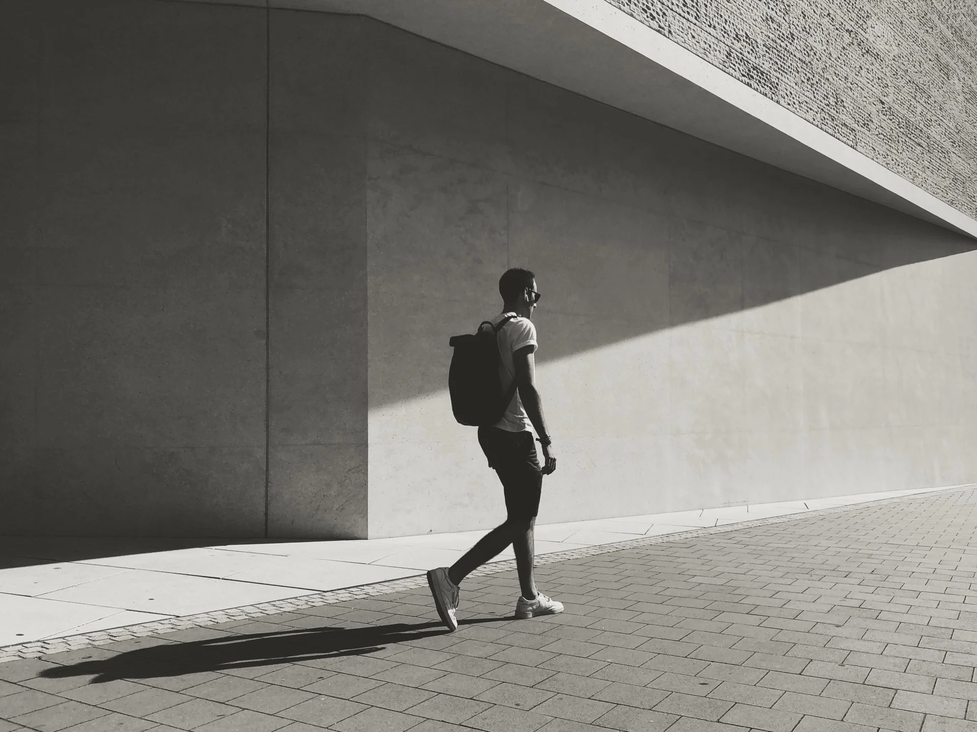 Young man walking on a city sidewalk, wearing sneakers, a white t-shirt, glasses, and a backpack, with a bold shadow cast on the wall behind him.